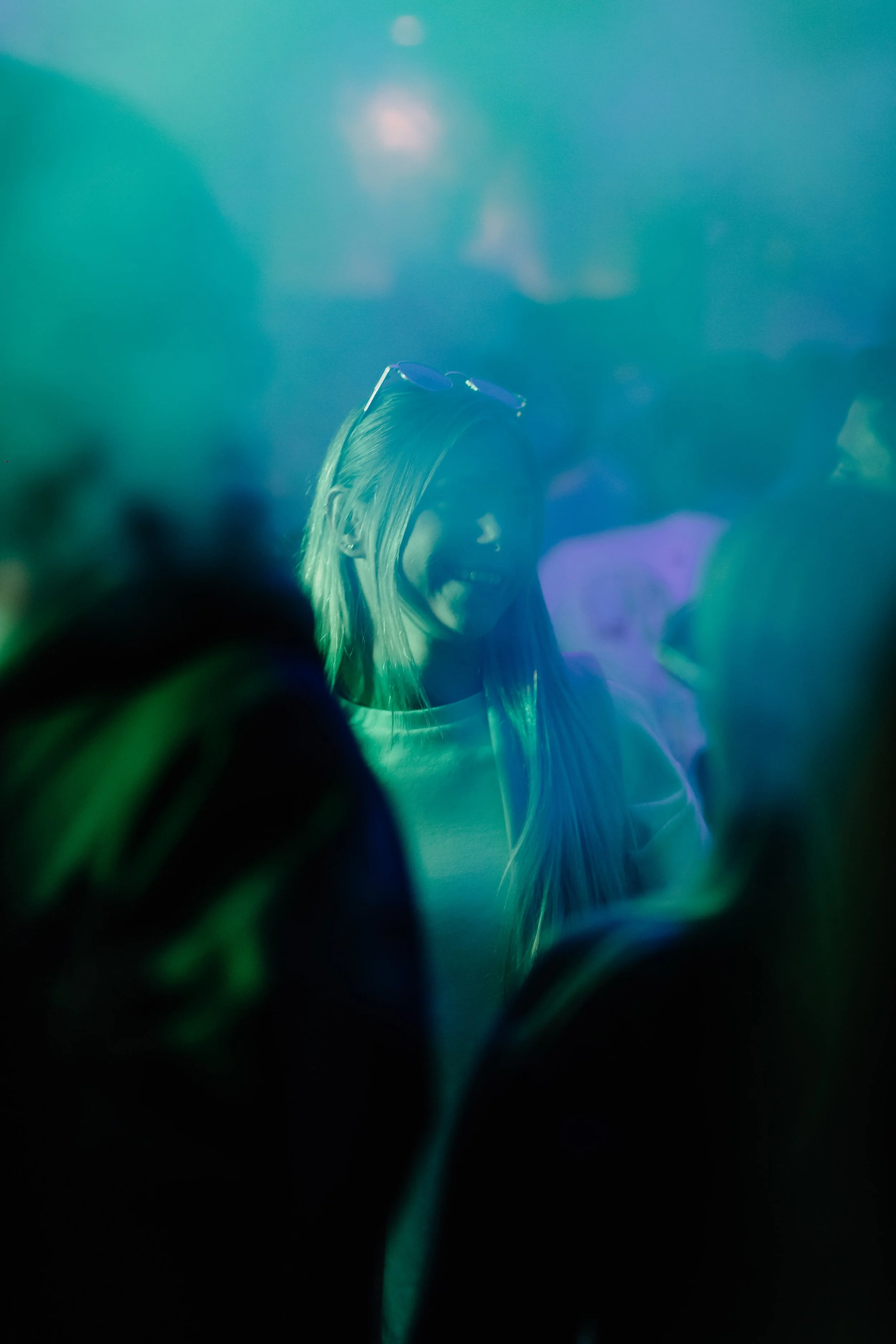 Een vrouw met lang blond haar en zonnebril op haar hoofd, omringd door mensen op een feestje of club, onder blauwe en groene verlichting.