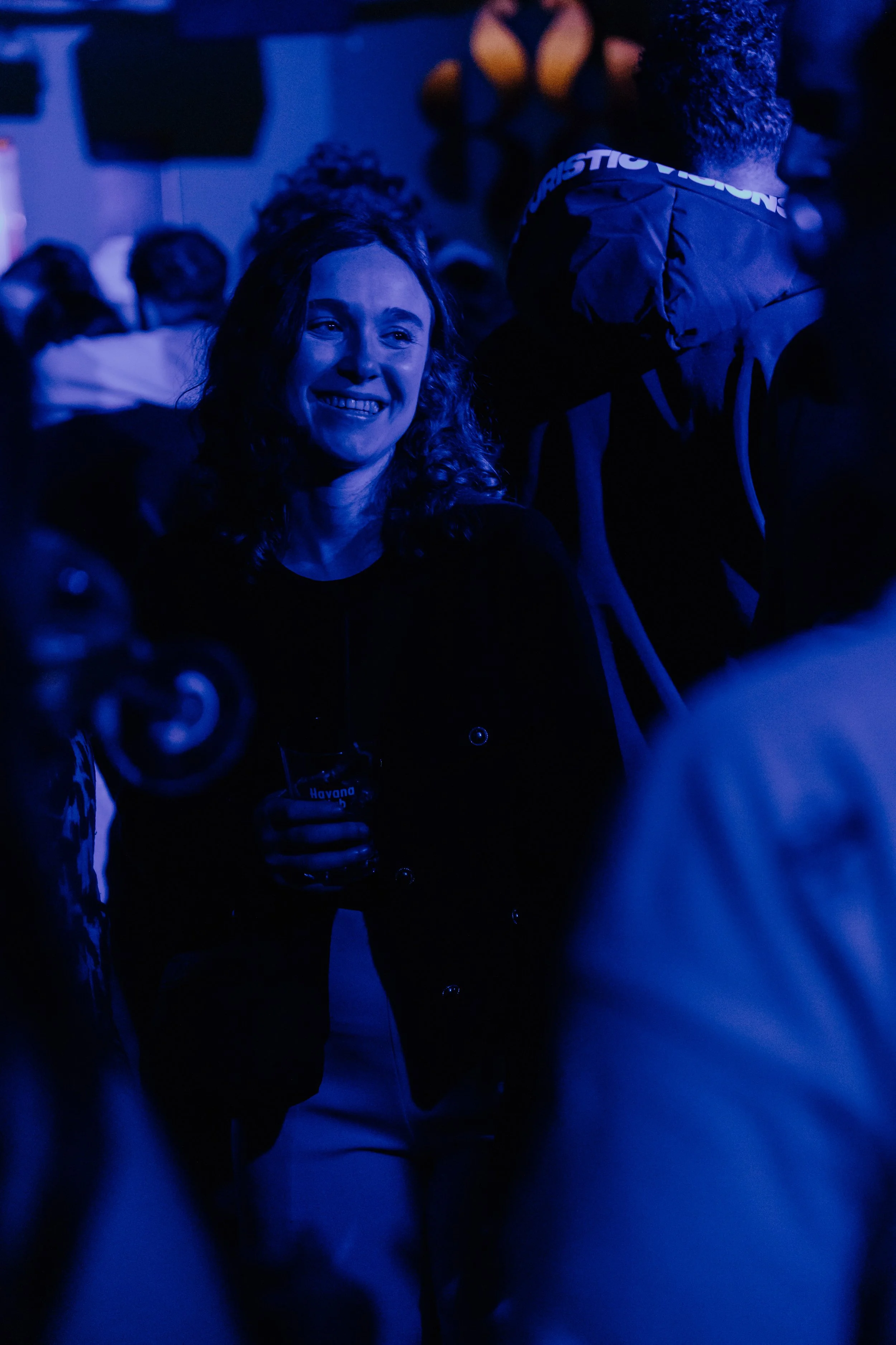 Vrouw met glimlach in een donker, blauw verlichte ruimte, mogelijk een bar of feest, houdt een glas in haar hand.