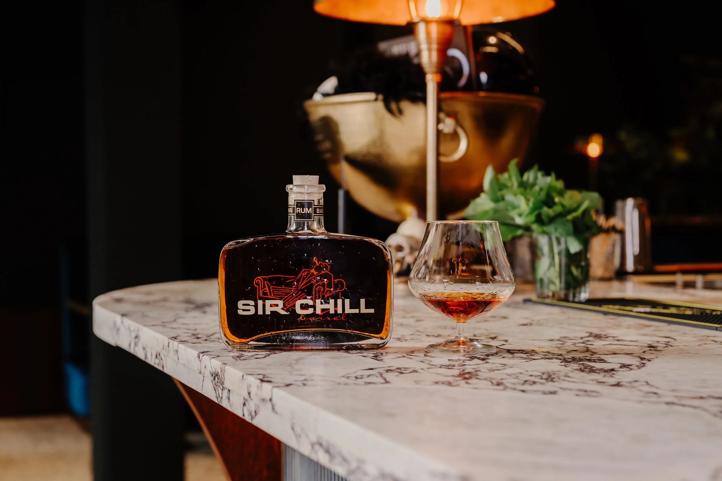Een fles Sir Chill likeur en een glas met roodgeel drankje op een marmeren bar, met een plant en een lamp op de achtergrond in een donkere setting.
