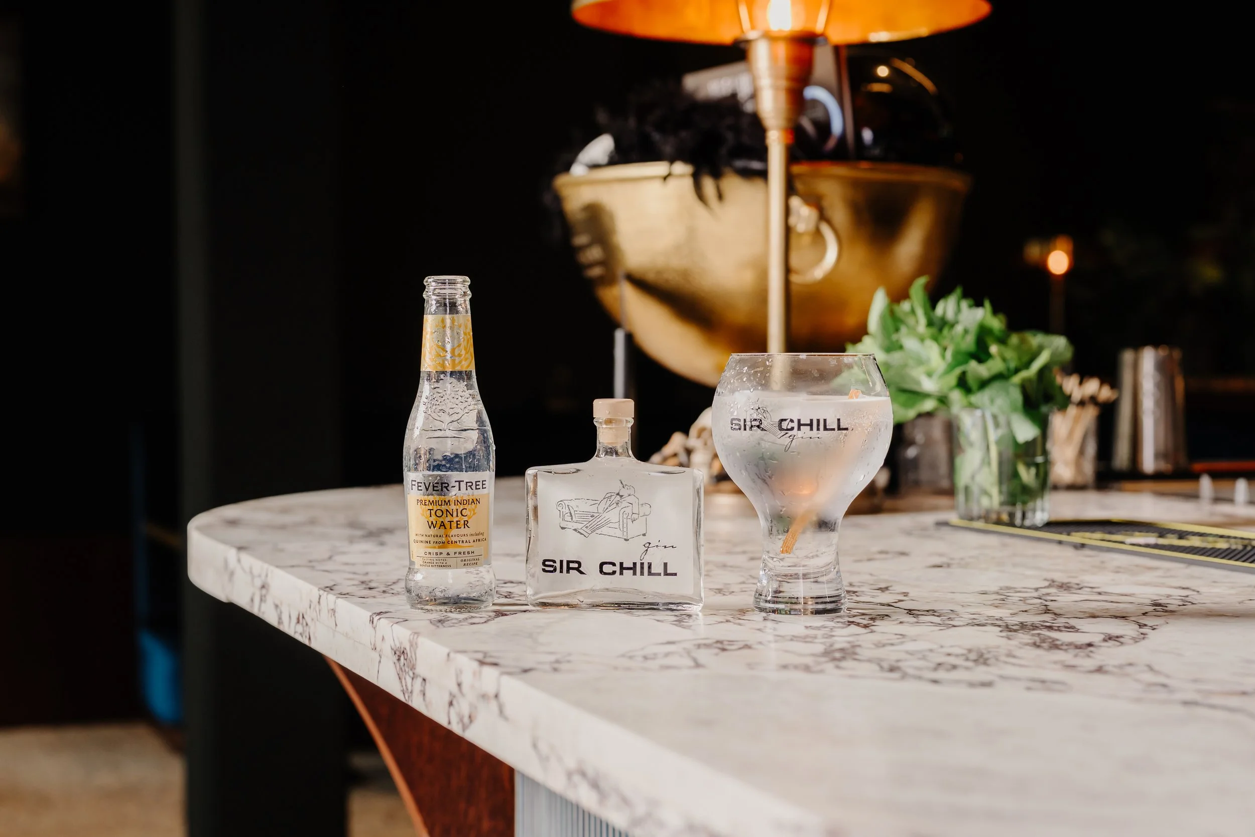 Een bar met een wit marmeren aanrecht, een fles tonic water, een fles gin en een glas met gin en ijs op de voorgrond. In de achtergrond bevindt zich een grote bronzen kom met een lamp erboven, en groene plant.