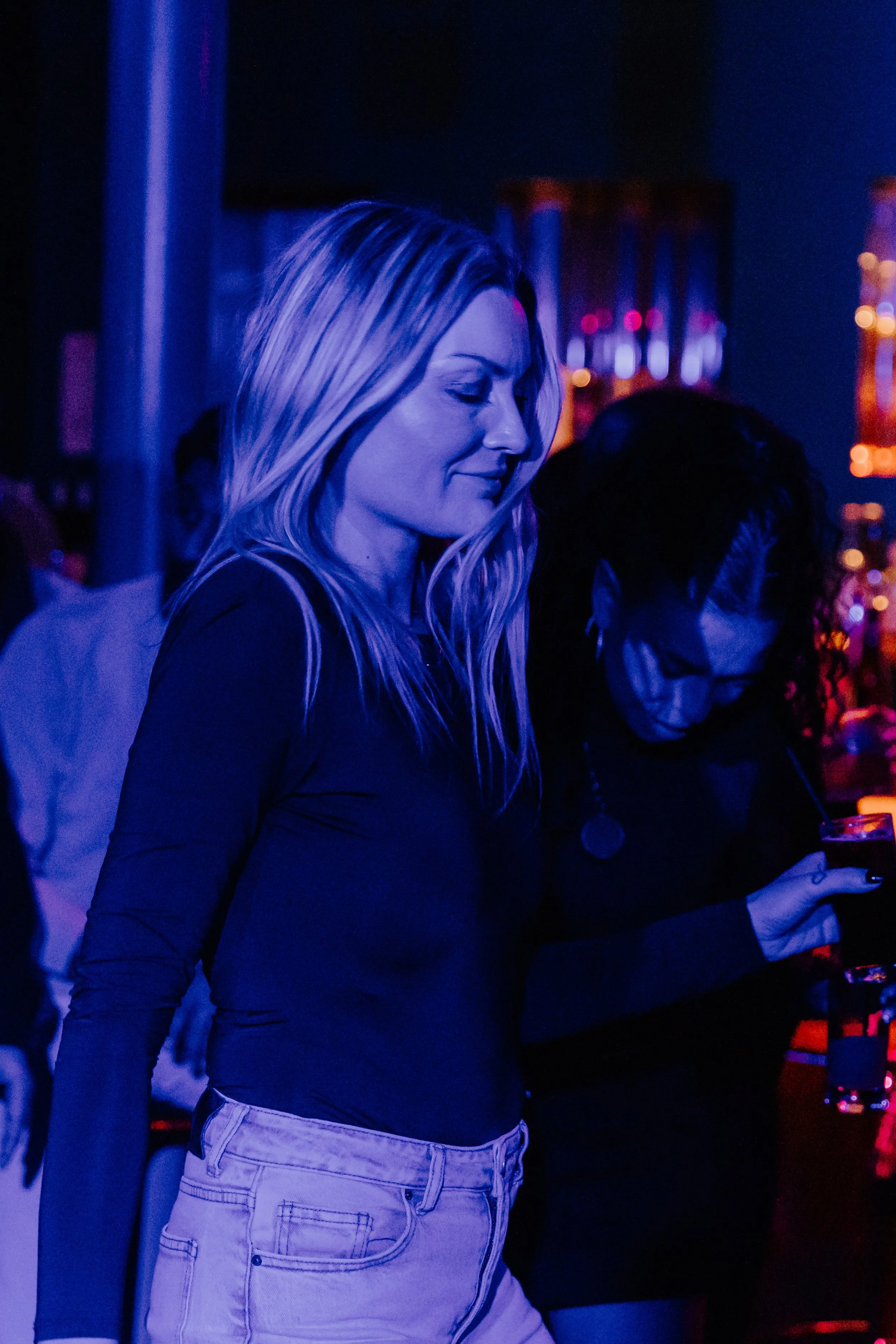 Twee vrouwen in een donker neon verlichte omgeving, een vrouw met lang blond haar en witte broek, en een vrouw met donker krullend haar die een drankje vasthoudt.