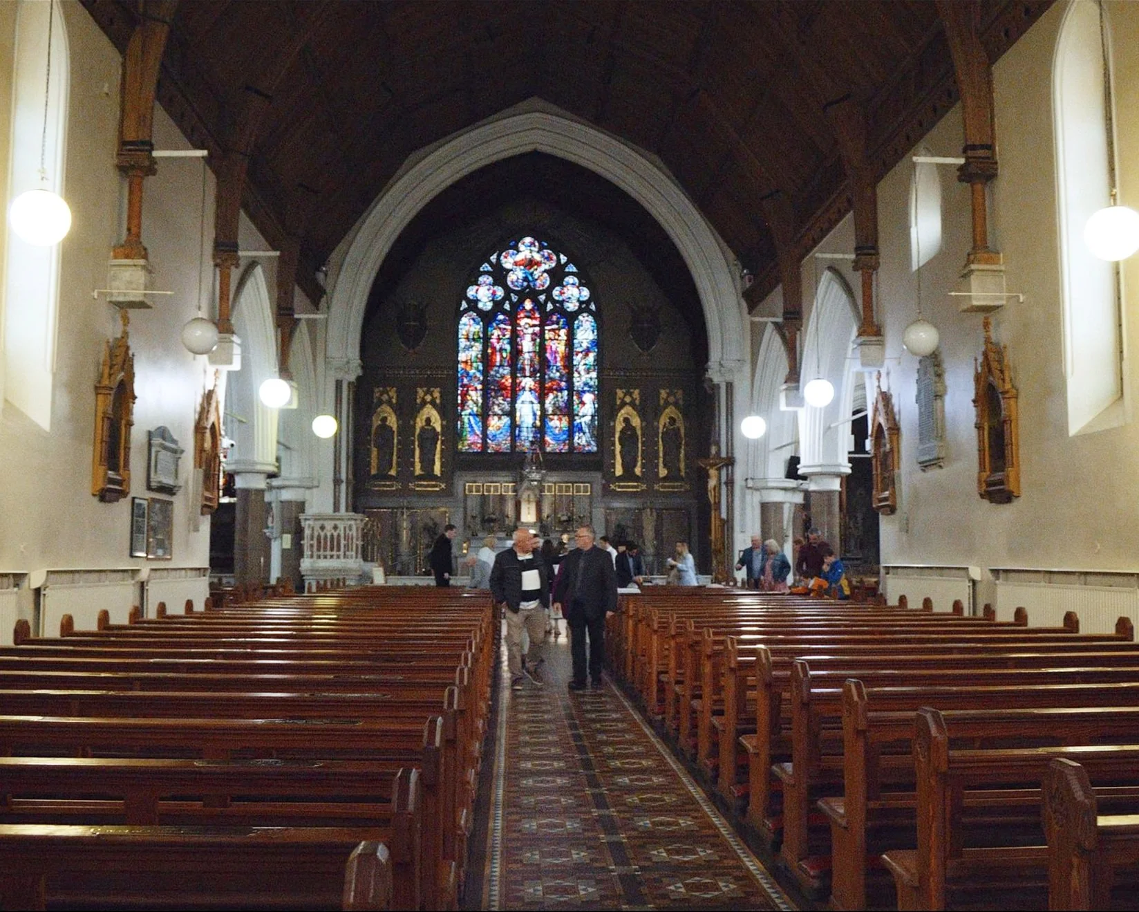 Interior of a church with wooden pews, stained glass window at the front, and people walking and talking.