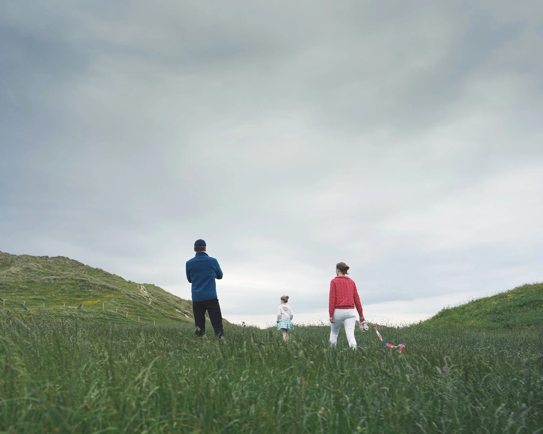 Three people, two children and one adult, walking on green grass in a hilly outdoor area under a cloudy sky, with one child holding a colorful ribbon.