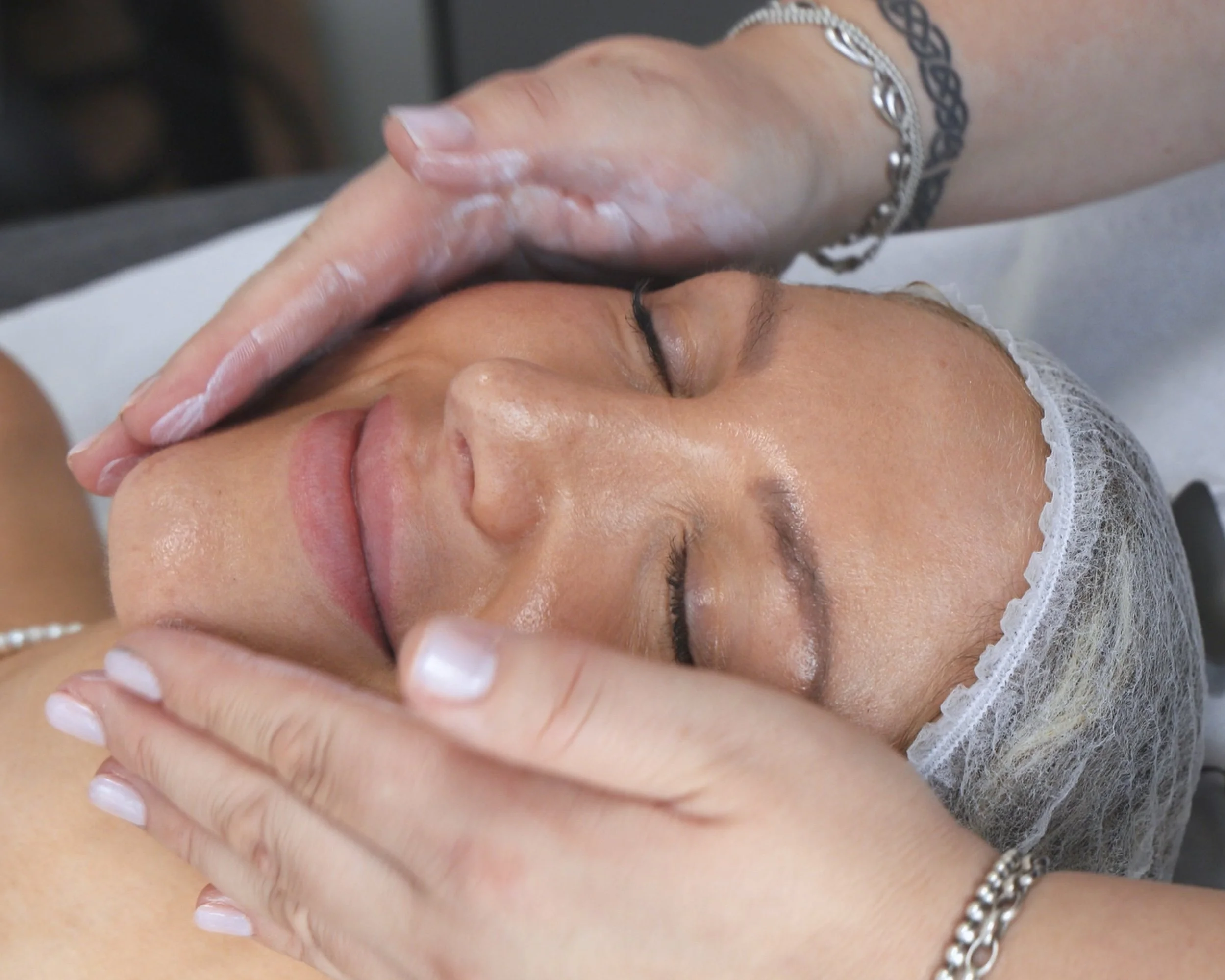 A woman receiving a facial massage at a spa or skincare clinic, lying down with eyes closed, wearing a hair cap and with hands gently massaging her face.