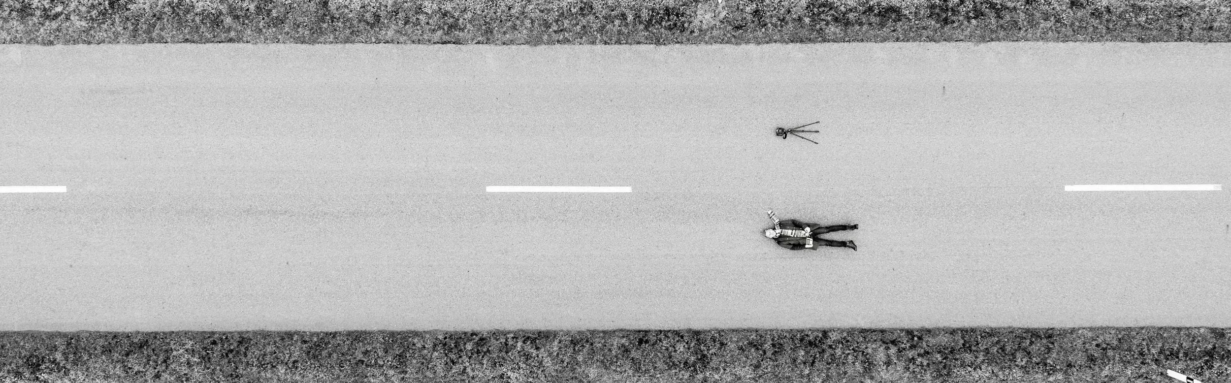 Black and white aerial view of a person walking along a two-lane road with a camera on a tripod nearby.