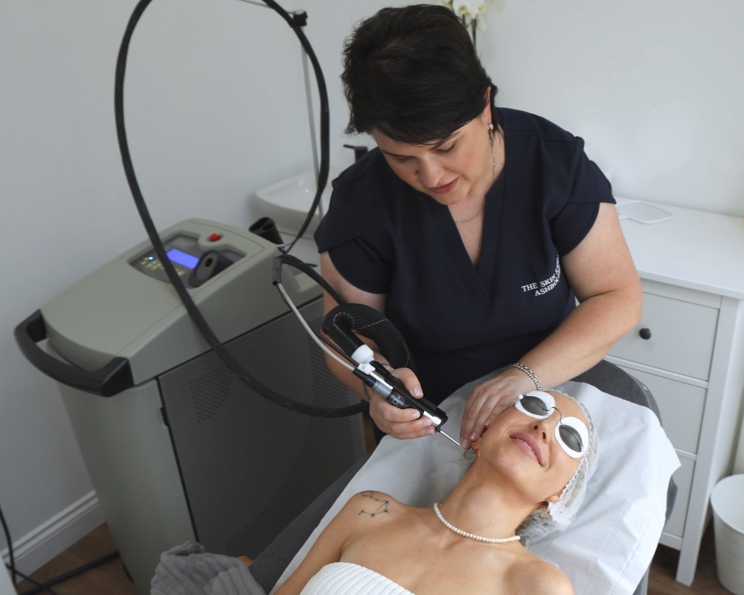 A woman lying on a treatment bed with protective eyewear undergoing a cosmetic procedure on her face, while a technician using a handheld device performs the procedure in a clinic or spa setting.