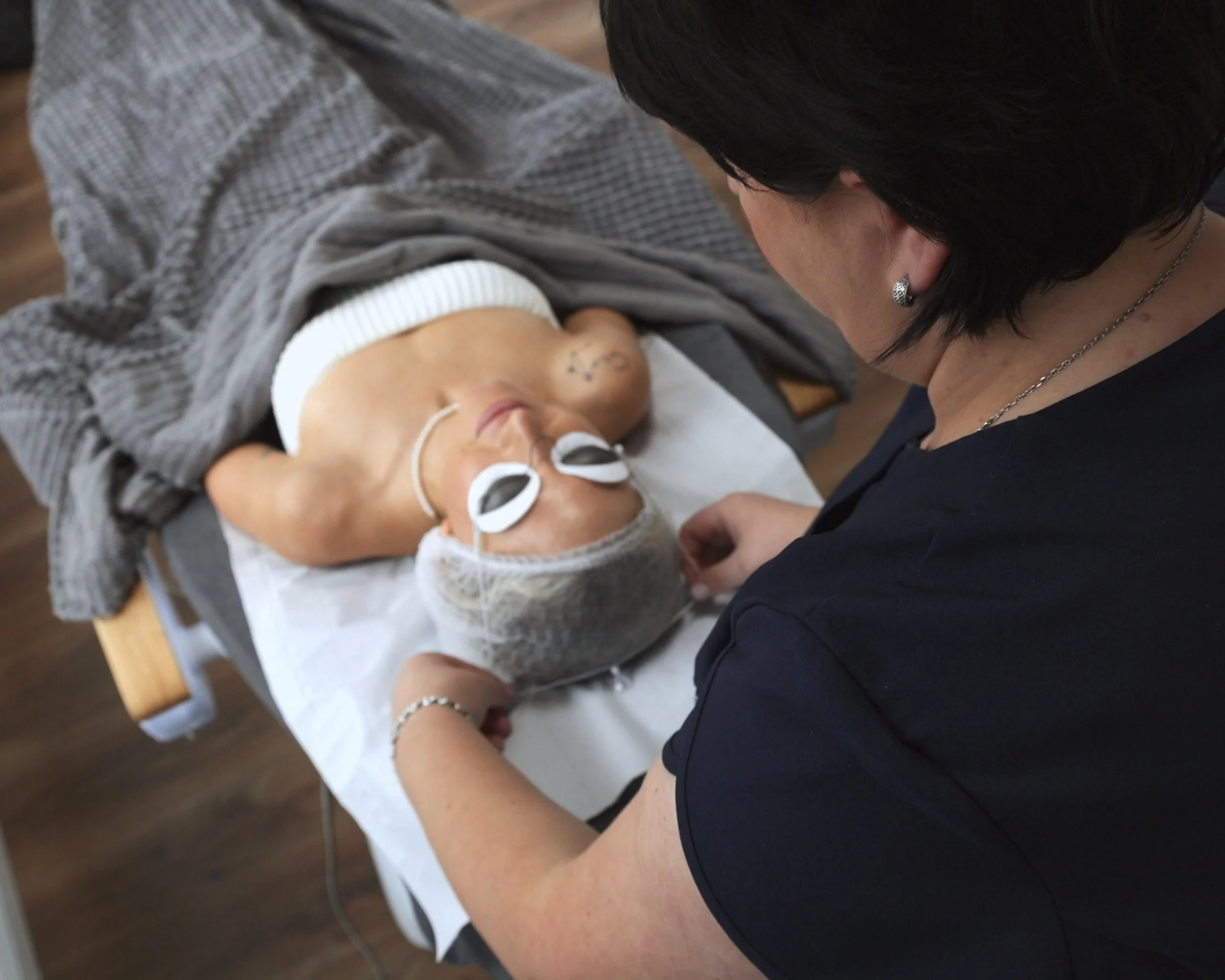 A woman is lying on a medical examination table with a blanket, while a healthcare professional attends to her, wearing protective gear and goggles, in a clinical setting.