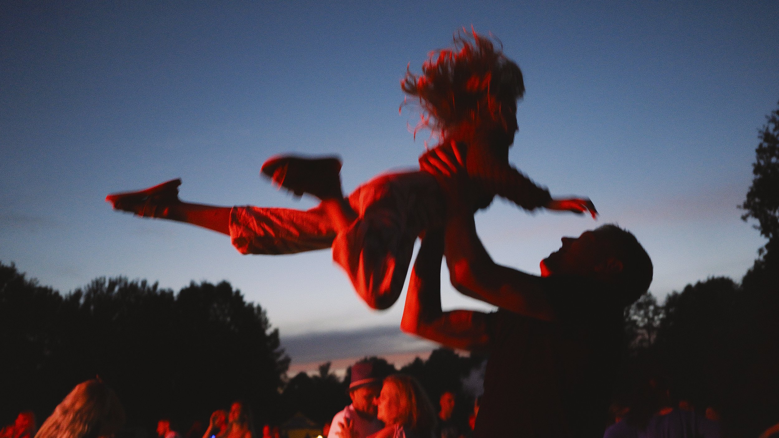 A silhouette of a person lifting another person in the air during sunset or dusk, with trees and a group of people in the background.