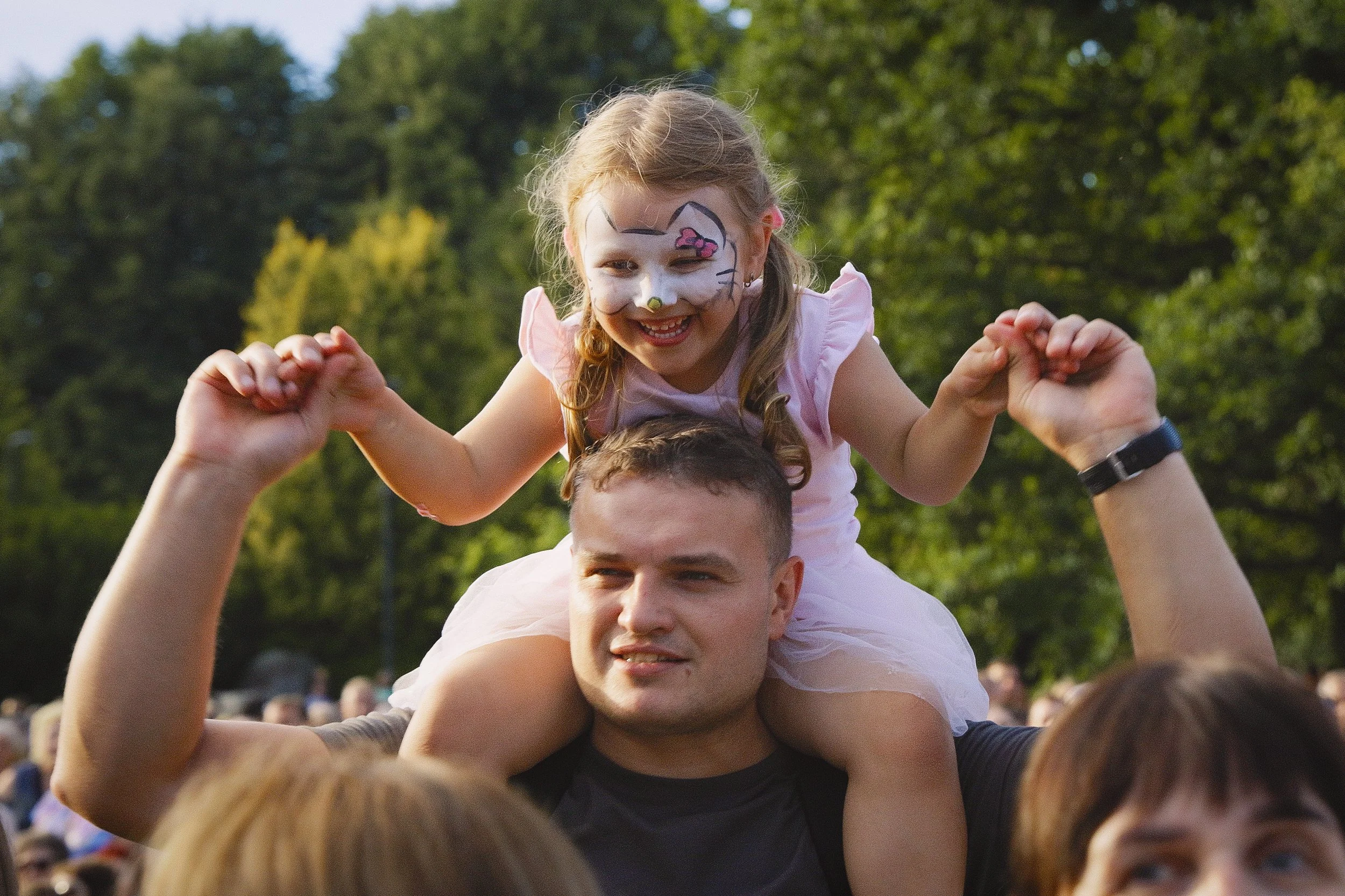 A young girl with face paint of a cat, smiling and sitting on a man's shoulders in a crowd outdoors, with trees in the background.