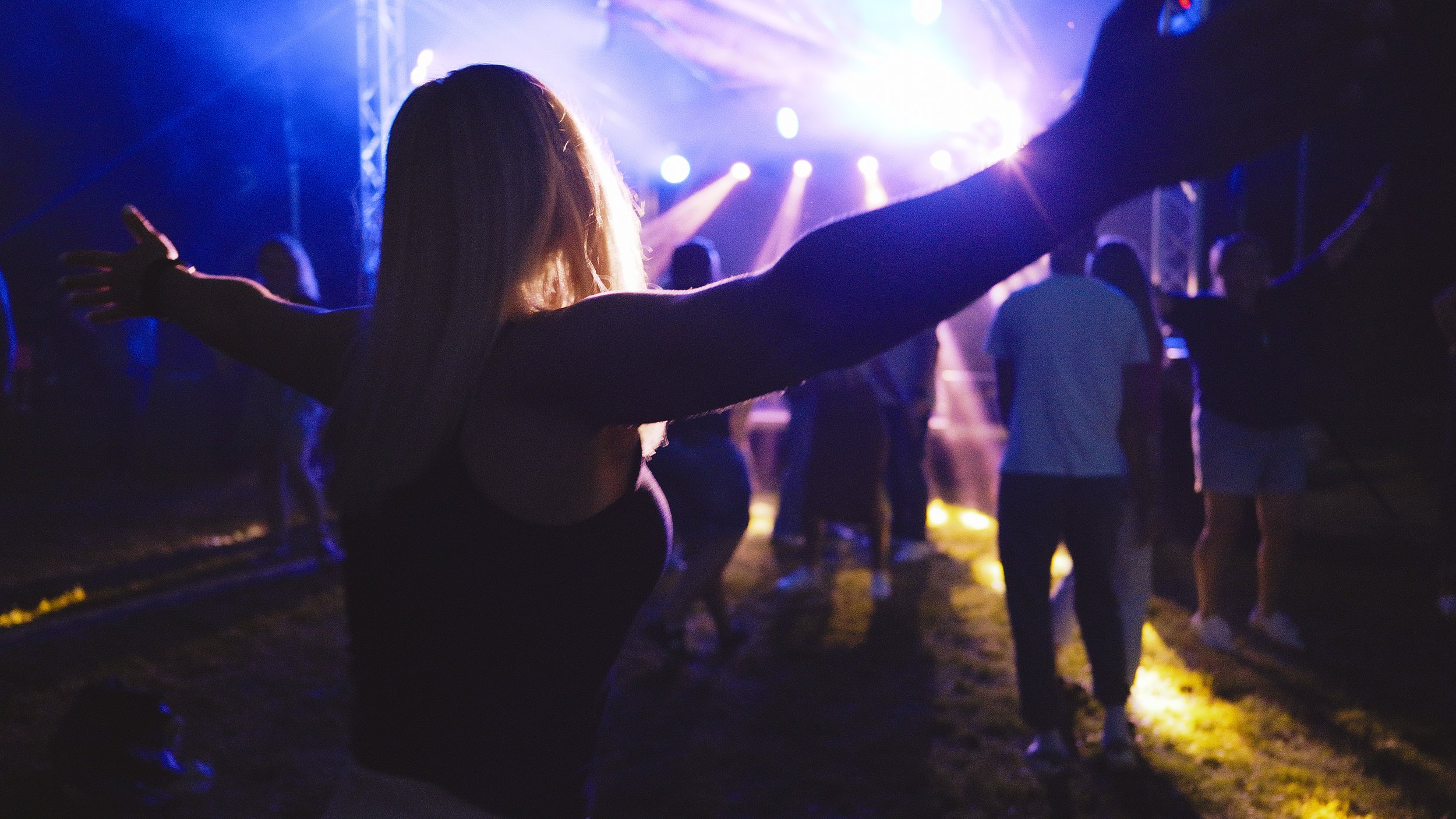 A woman with arms outstretched at a concert or event, surrounded by people with colorful stage lighting and a dark background.