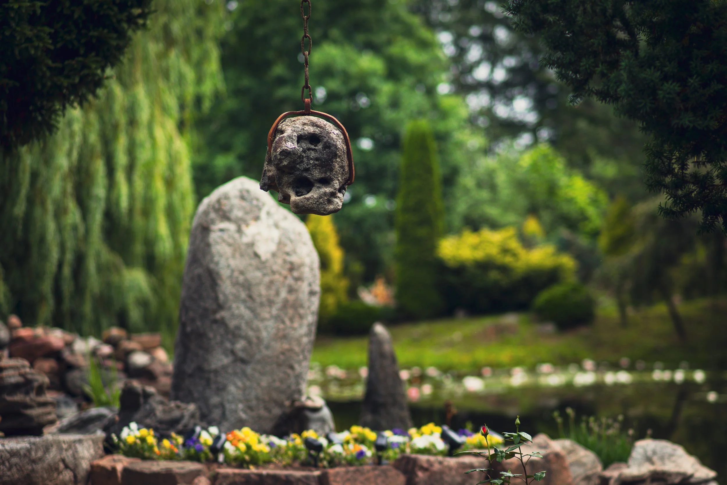 A stone sculpture of a skull with a chain hanging from above, placed on a rock surrounded by colorful flowers, in a lush garden with trees and greenery.