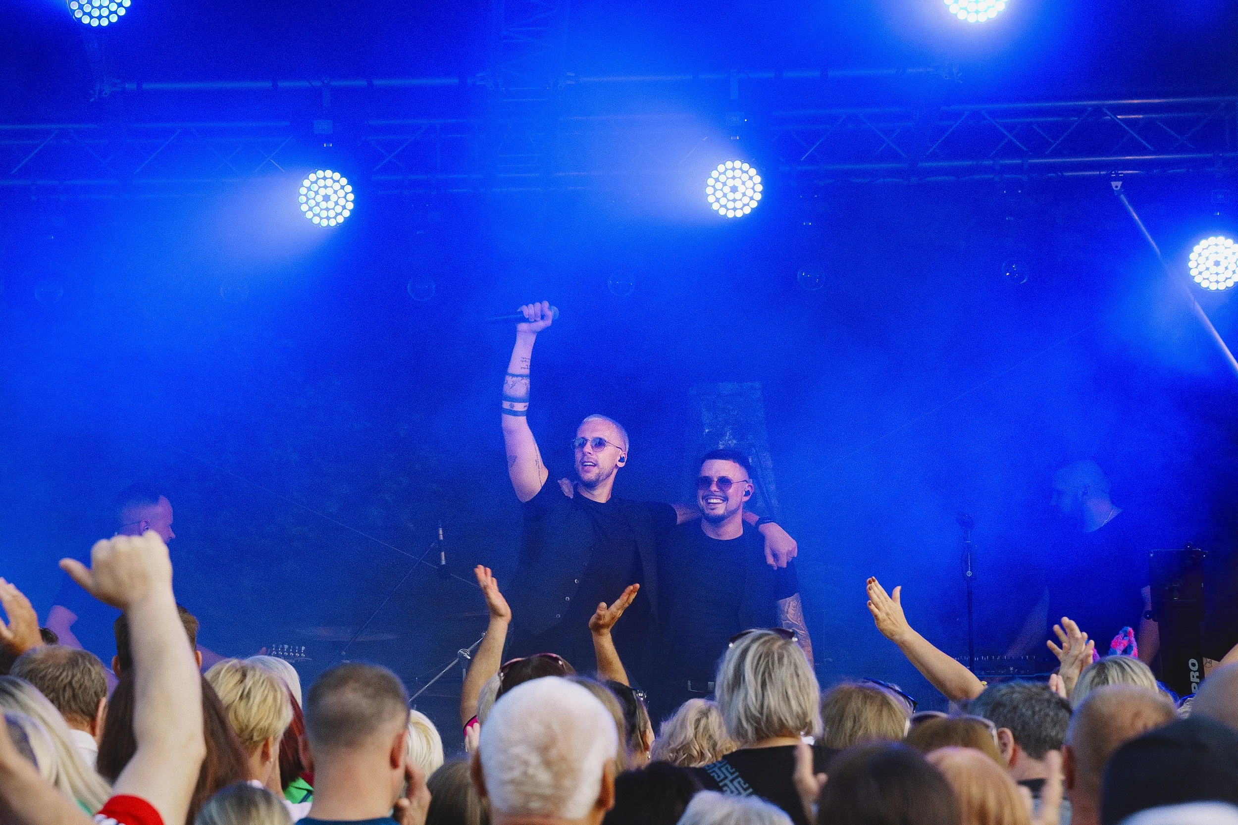 Two men on stage, one holding a microphone, with a crowd in front of them at a concert or event under blue lighting.