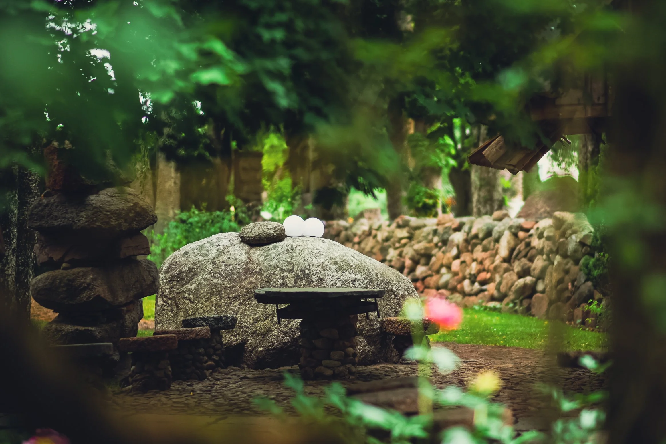 A tranquil garden scene with a large rock, smaller stacked rocks, and two white orbs resting on the large rock. Surrounding greenery and a stone path lead through the garden, which is enclosed by a rock wall.