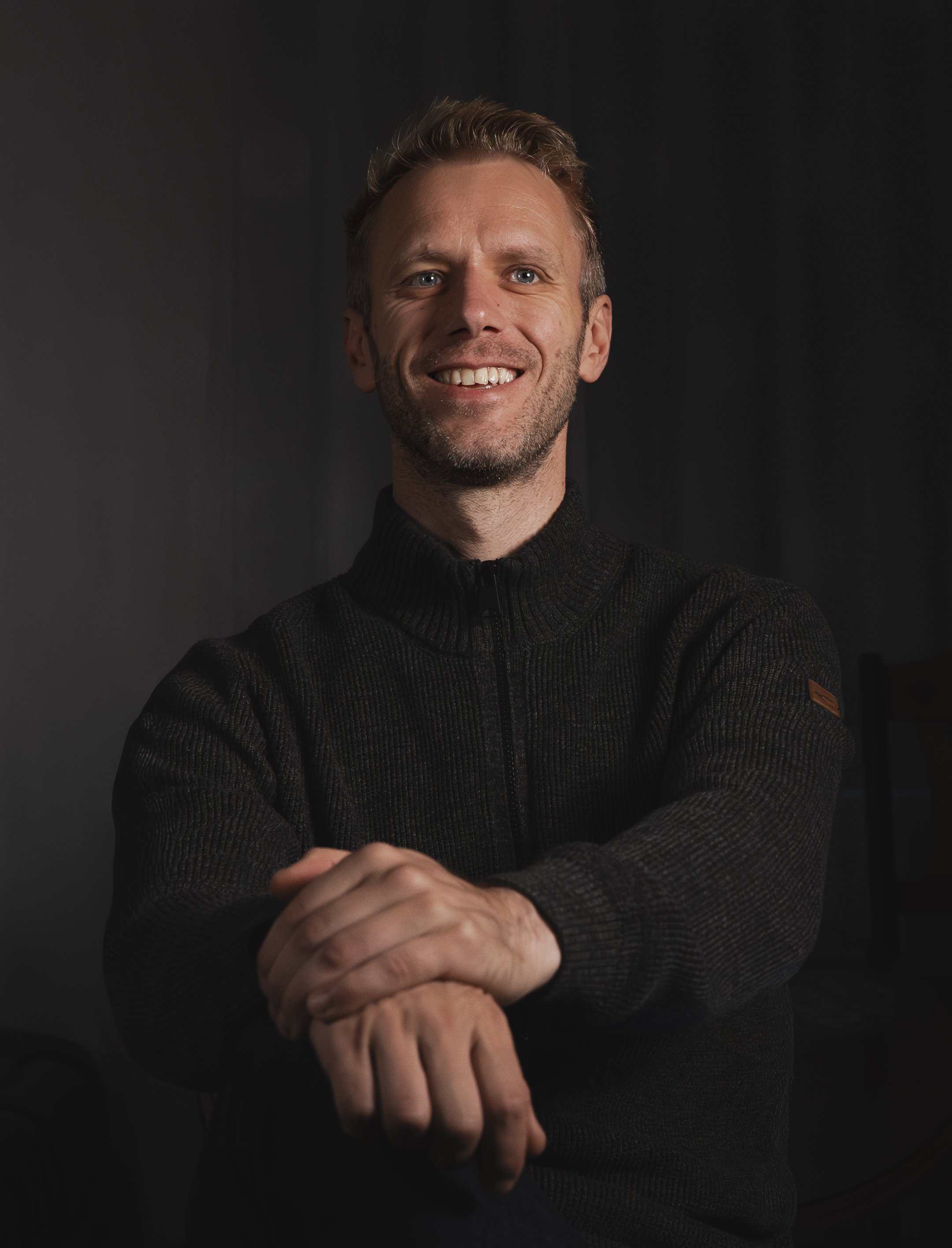vyras, trumpais šviesiais plaukais ir barzda, dėvintis juodą megztinį, sėdintis prieš tamsų foną.
