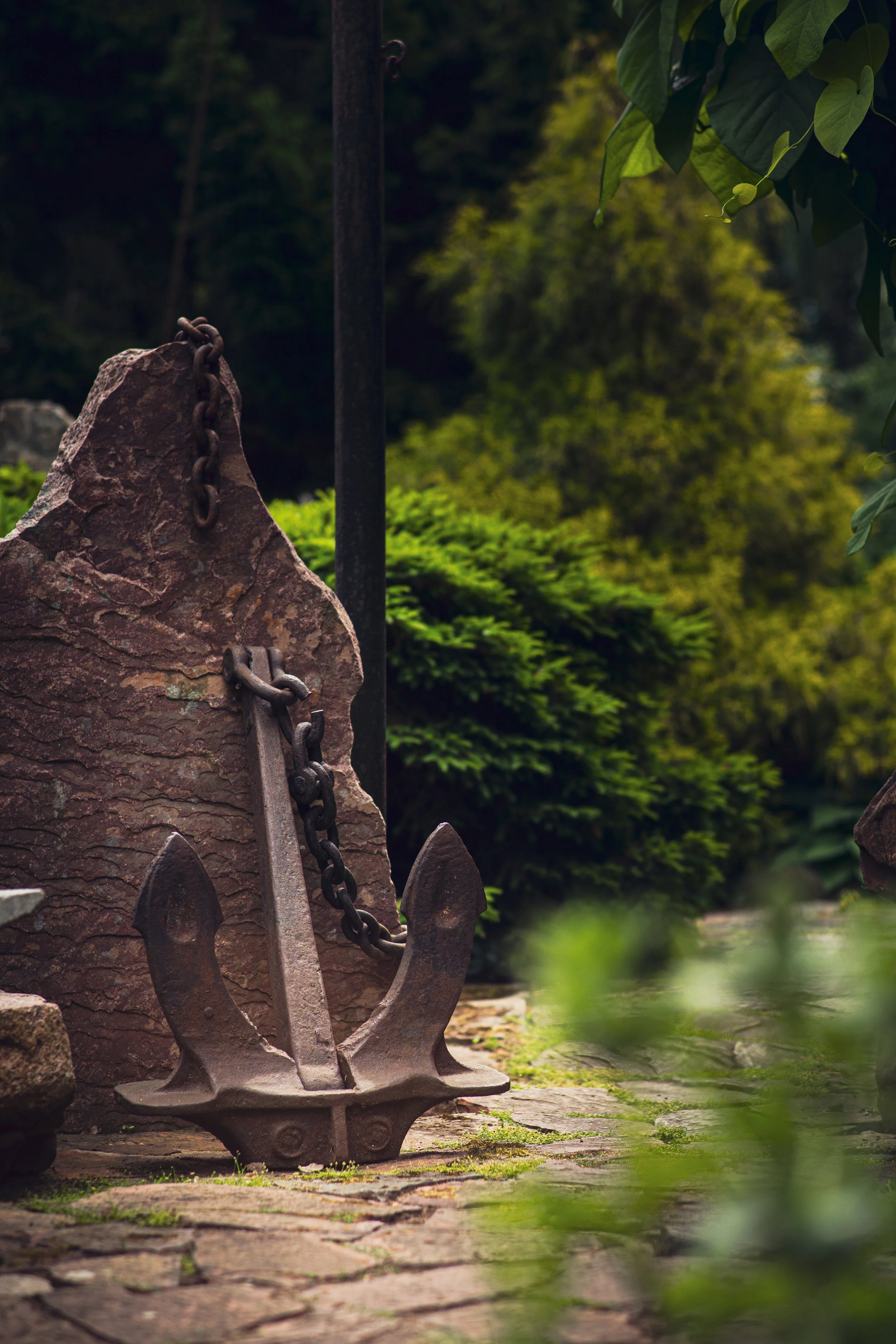 A rusty anchor leaning against a large rock in a garden with green bushes and trees in the background.