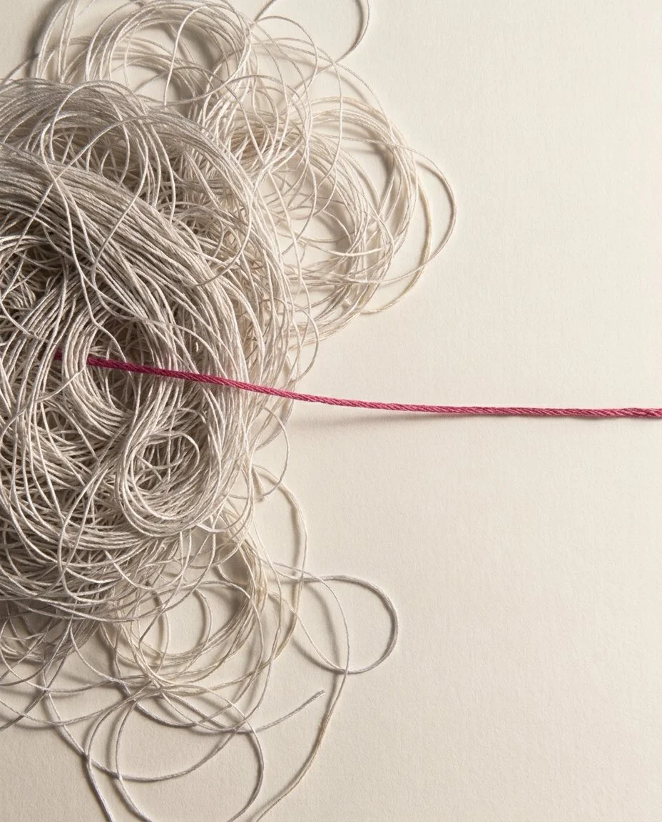 A dense tangle of pale threads with a single taut magenta thread emerging from the centre and pulled clear across a warm off-white surface.