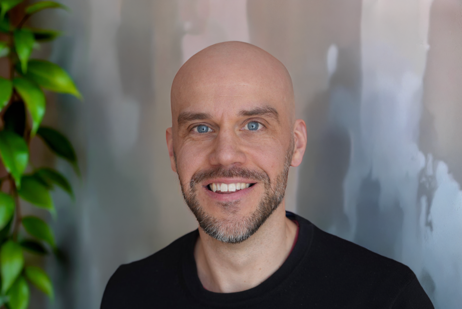 A smiling man (same guy as before, what could he possibly want, putting up a second photo of himself?) with a bald head and blue eyes, wearing a black top, standing against a background with a metallic surface and some green leaves on the left side.