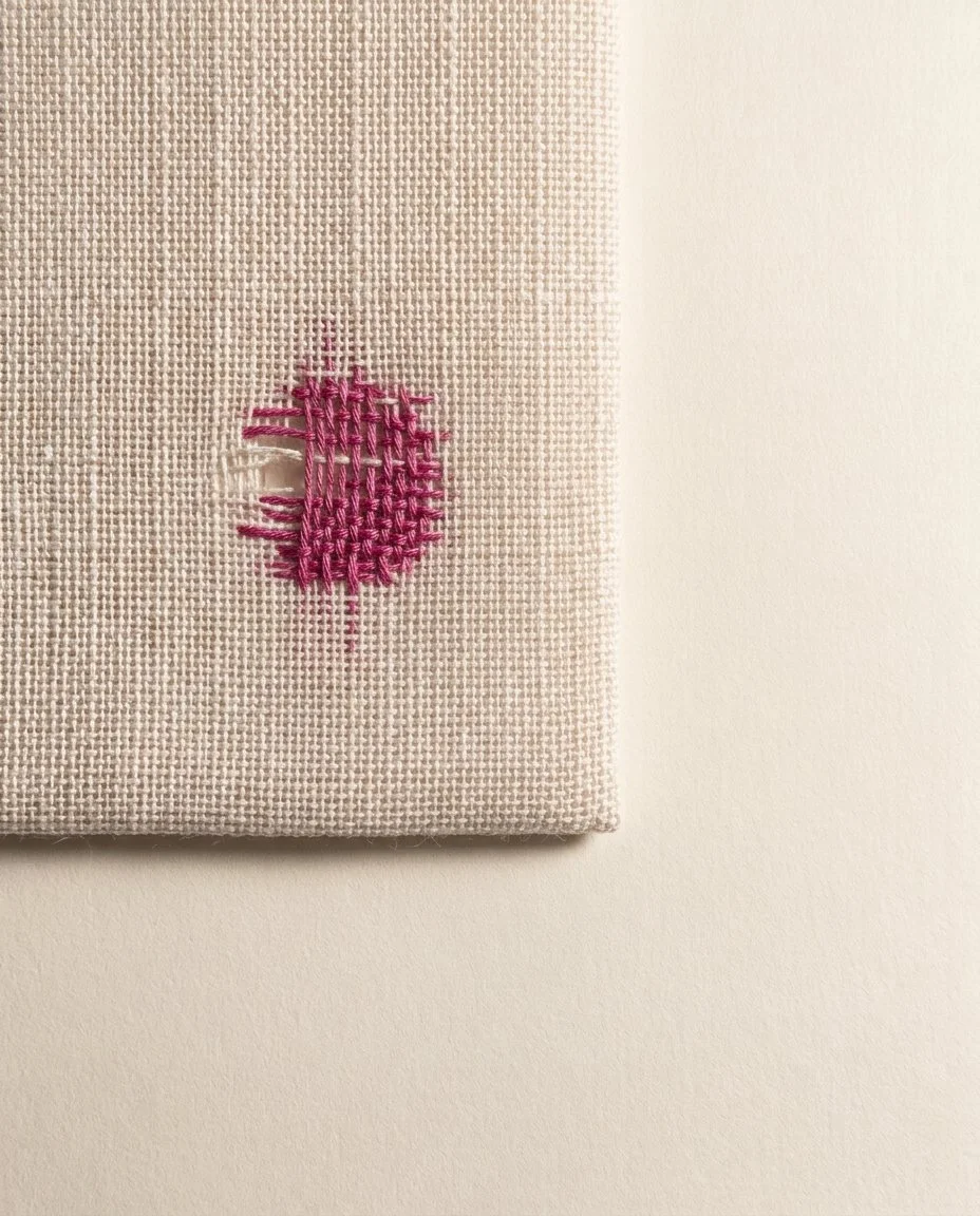 Pale woven linen with a gap darned closed by a patch of magenta thread woven over and under across the break.