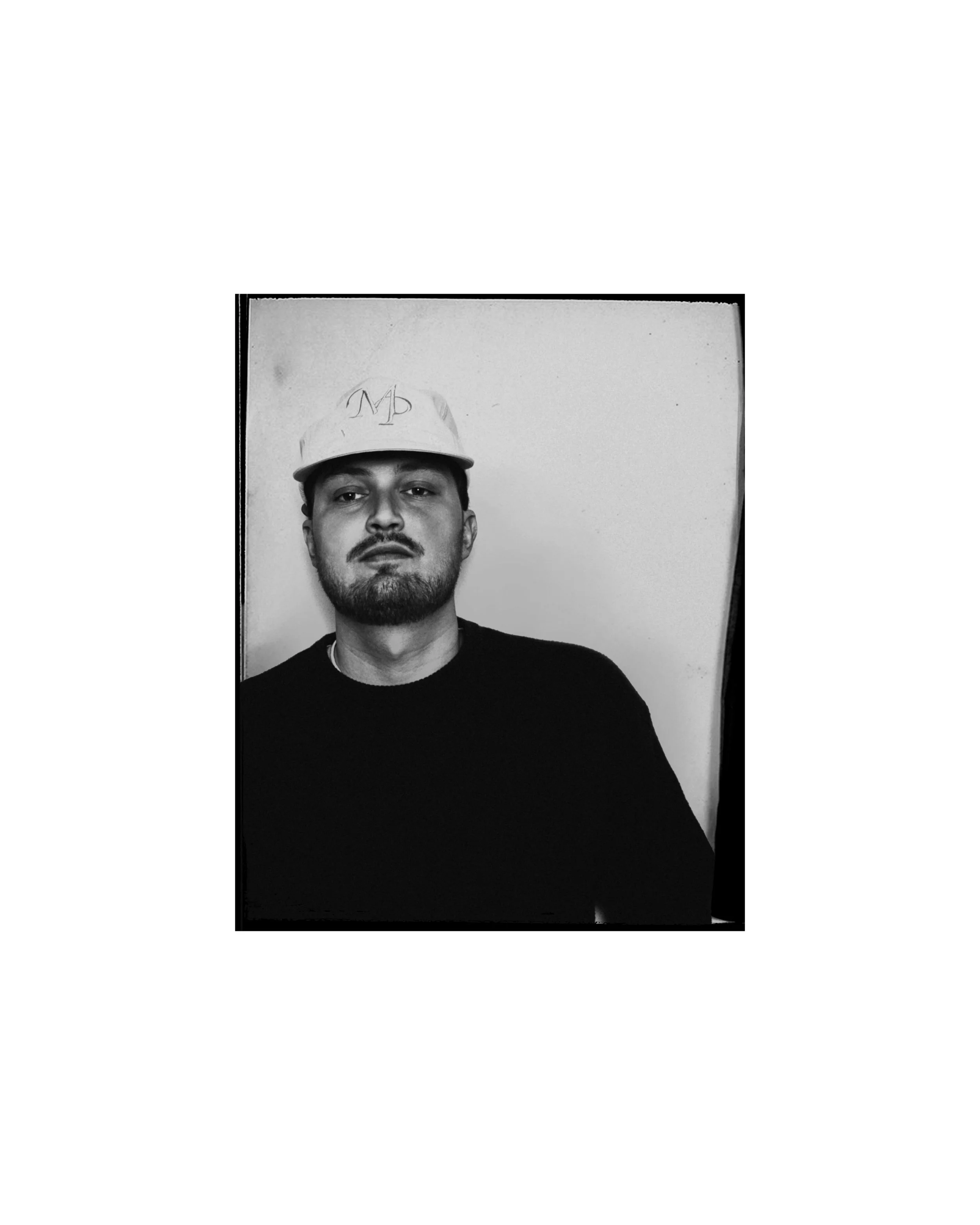 Black and white portrait of a man wearing a baseball cap and black shirt, looking directly at the camera, against a plain background.