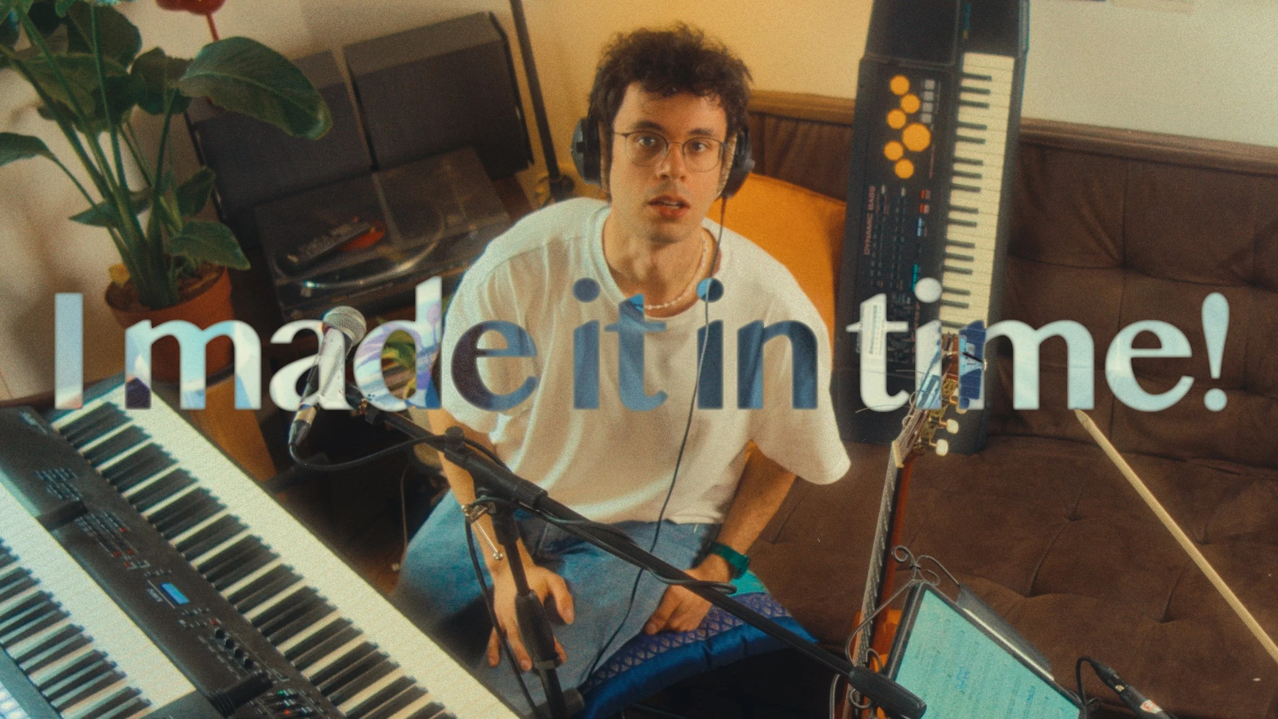 A man with curly hair and glasses wearing headphones, sitting in a room filled with musical equipment, including keyboards and a microphone, with a large text overlay saying 'I made it in time!'