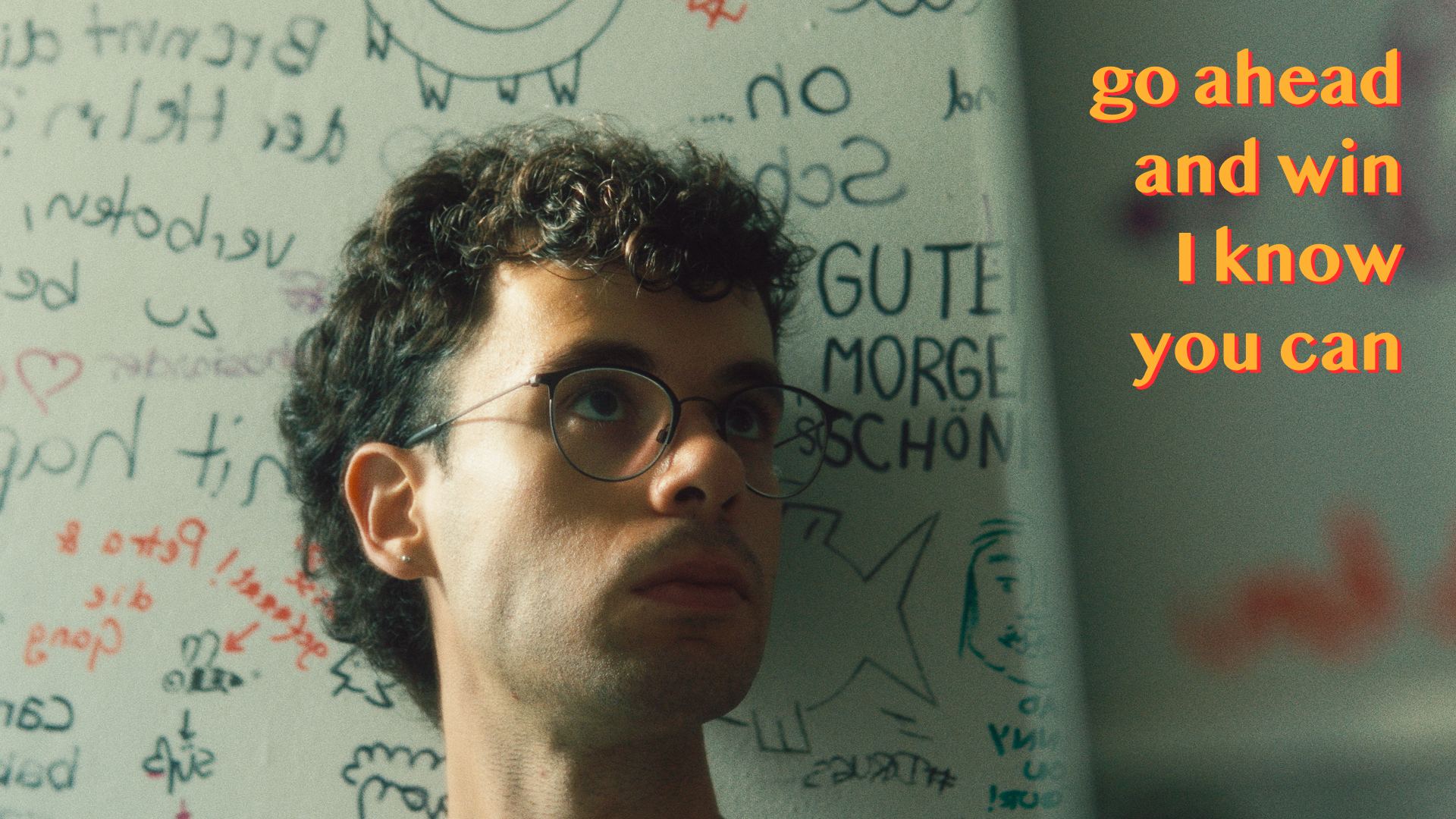 Person with glasses in front of a graffiti-covered wall, with words "go ahead and win I know you can" in bold orange text.
