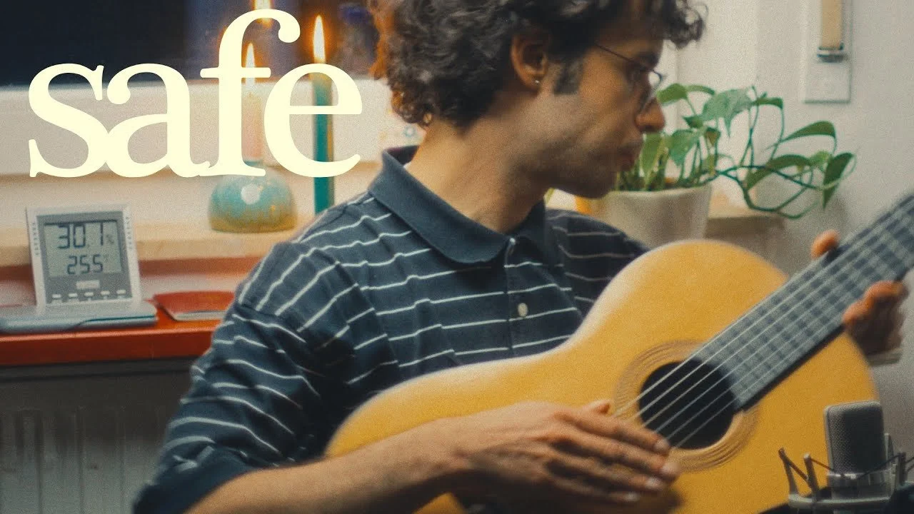 A person with curly hair wearing glasses and a black and white striped shirt is playing an acoustic guitar. The background features a potted plant, a digital clock displaying 30°C and 255, and a lamp, with the word 'safe' overlayed on the image.