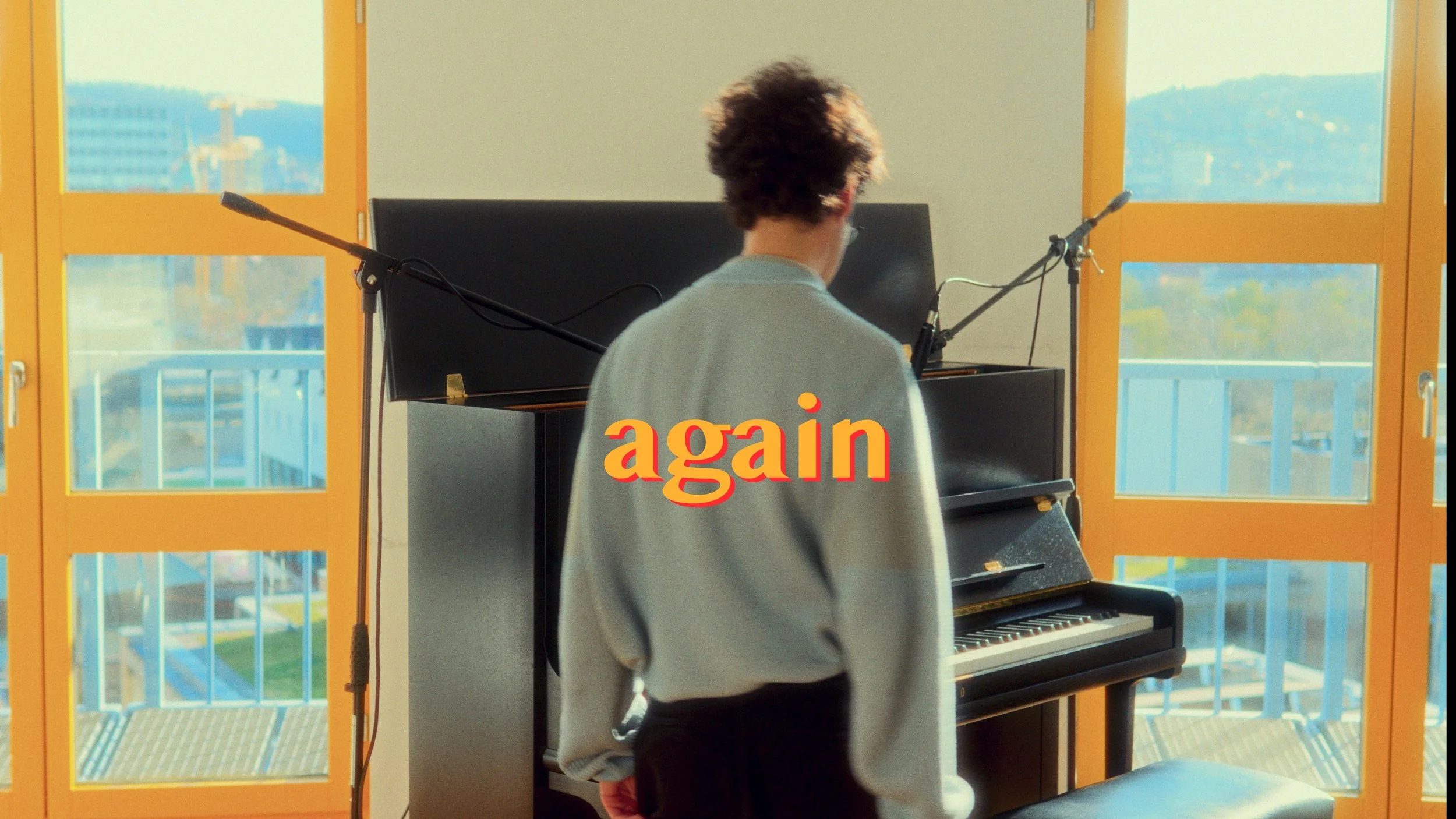 A person with curly hair in a light-colored shirt standing in front of a black upright piano with microphones pointed at it, in a room with large yellow-framed windows showing a cityscape outside. The word "again" overlays the image.