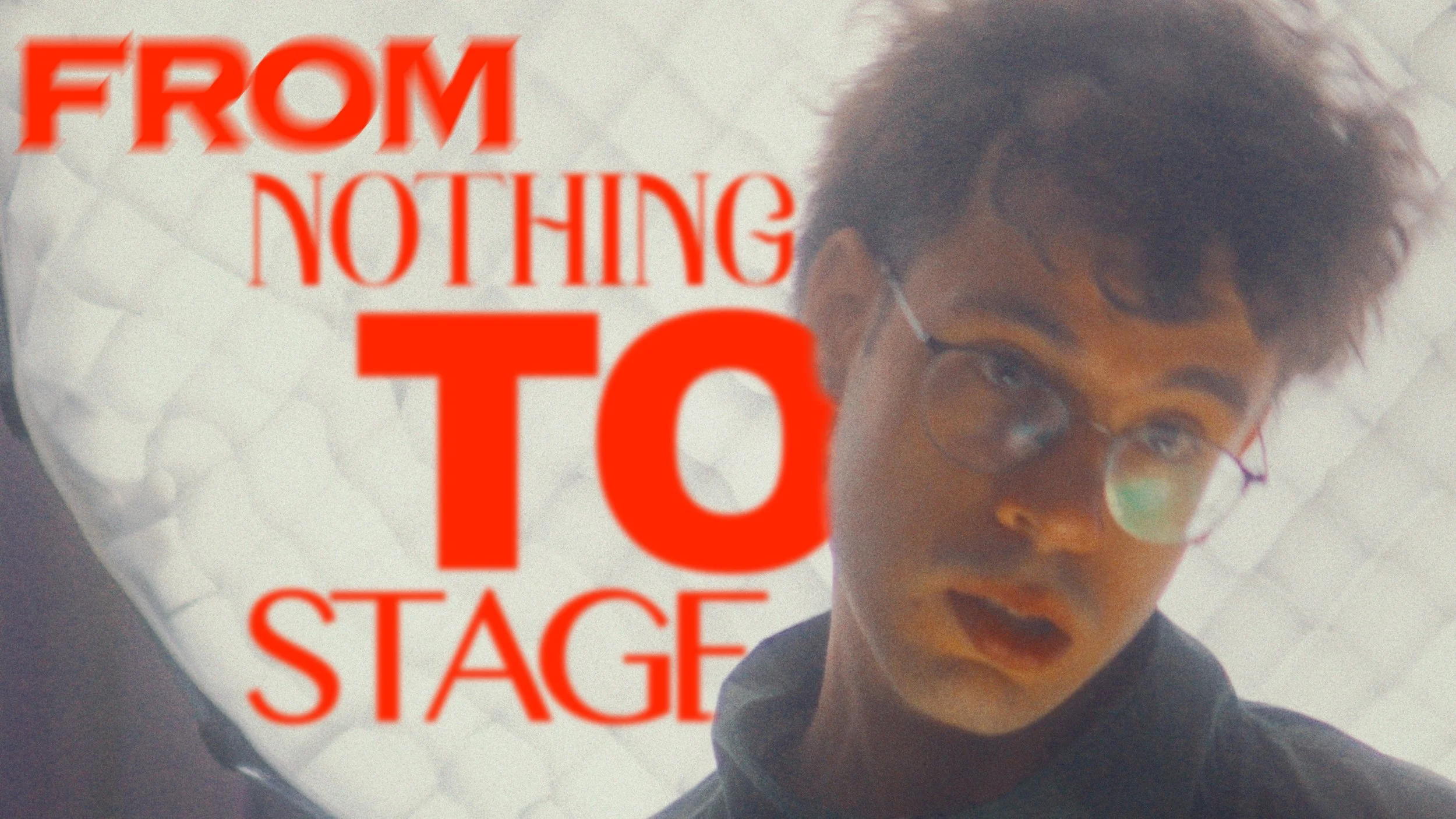 Young man with glasses and curly hair looking at the camera in front of a white background with red text reading 'FROM NOTHING TO STAGE'.