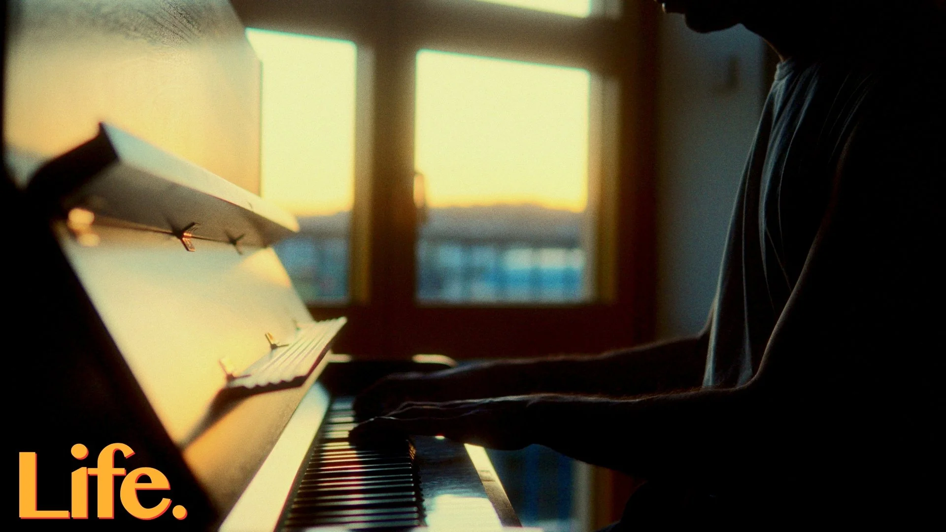 Person playing piano during sunset, with sunlight visible through window in background, and the word 'Life.' in orange text at the bottom left corner.