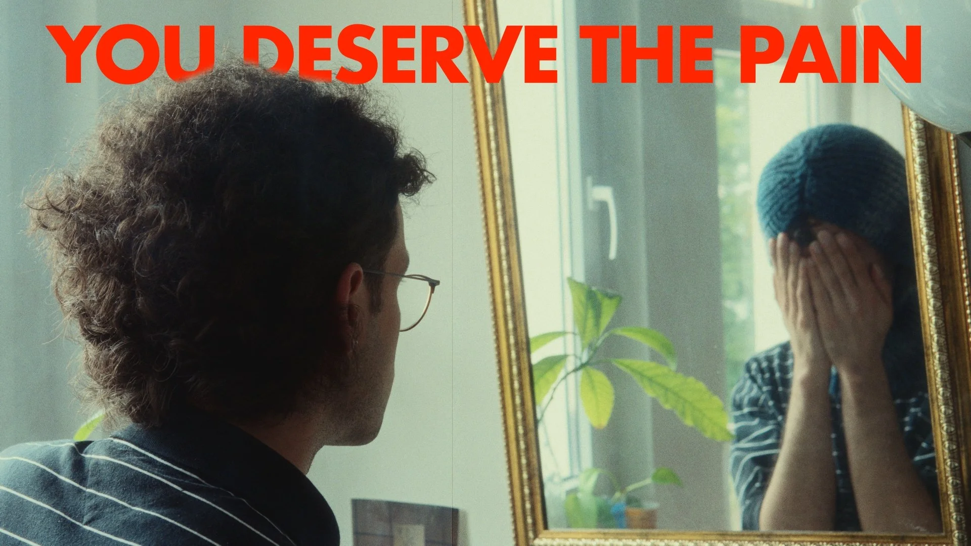 A woman covering her face with her hands looking at her reflection in a mirror, while a man with glasses looks on, with the text 'YOU DESERVE THE PAIN' in red at the top.