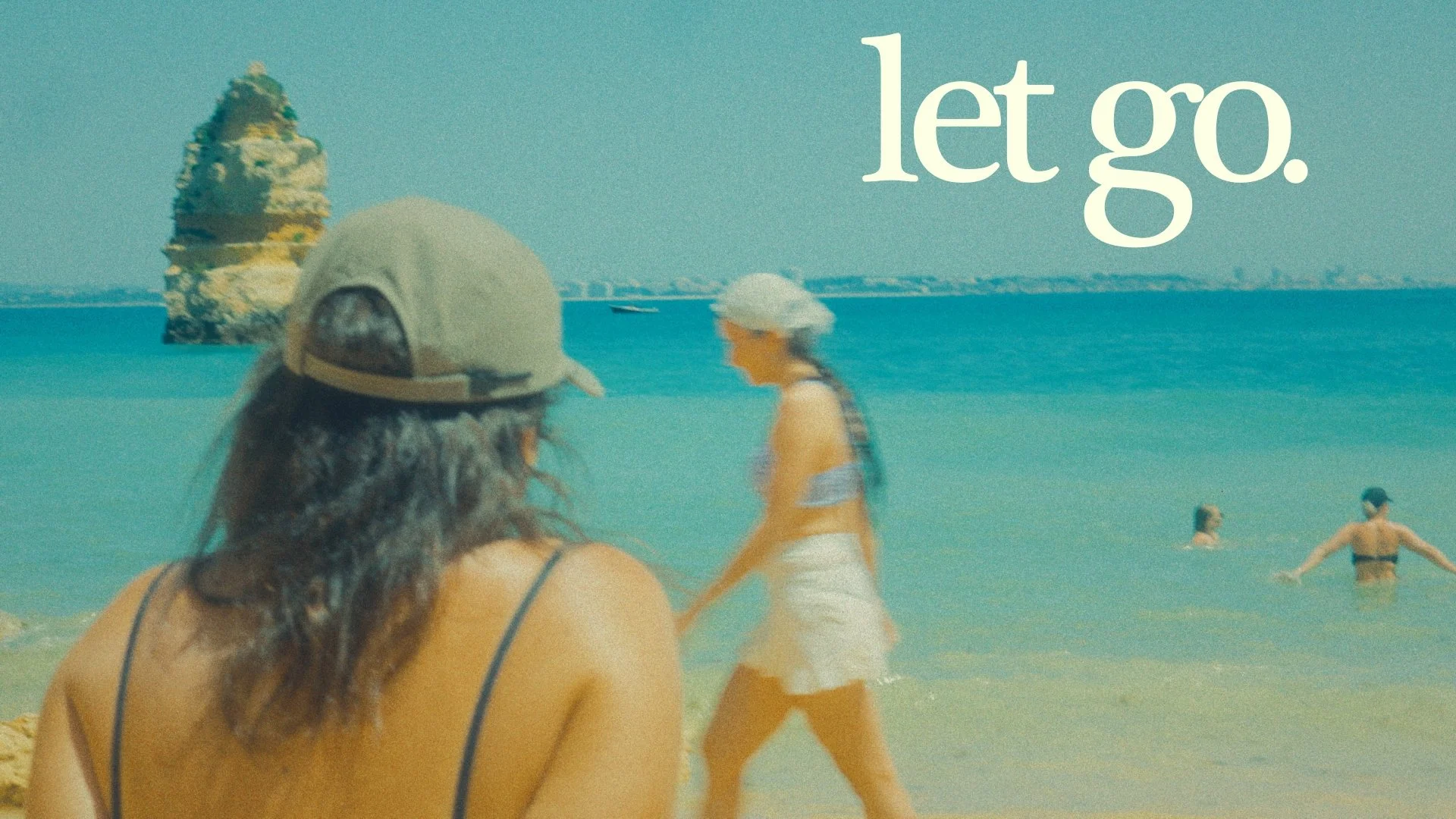 People relaxing and playing at the beach with a large rock or island in the background, under a clear blue sky, with the text 'let go.' in the upper right corner.