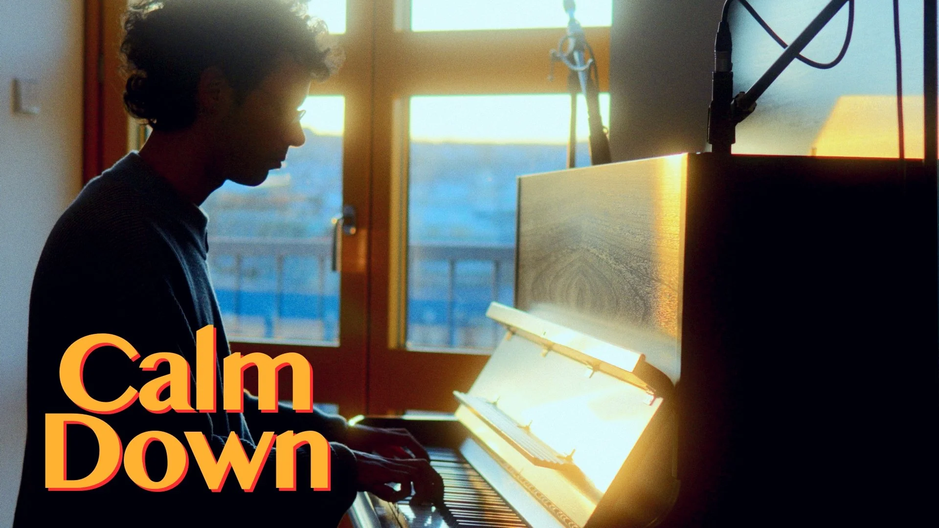 A person with curly hair playing a piano in front of a window during sunset, with the words 'Calm Down' in bold orange and yellow text in the lower left corner.