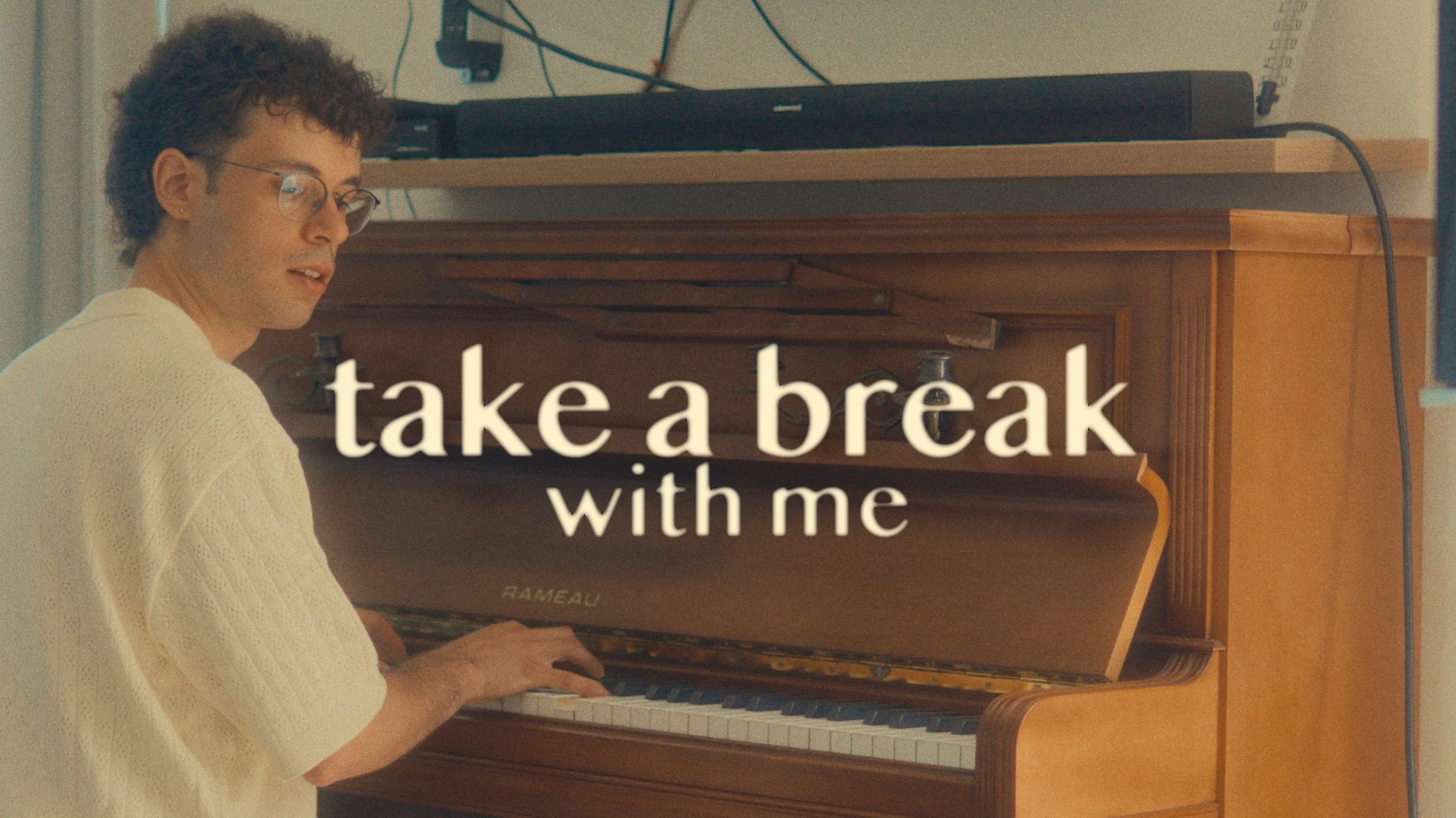 A person with glasses and curly hair playing a brown upright piano with the text 'take a break with me' overlayed.