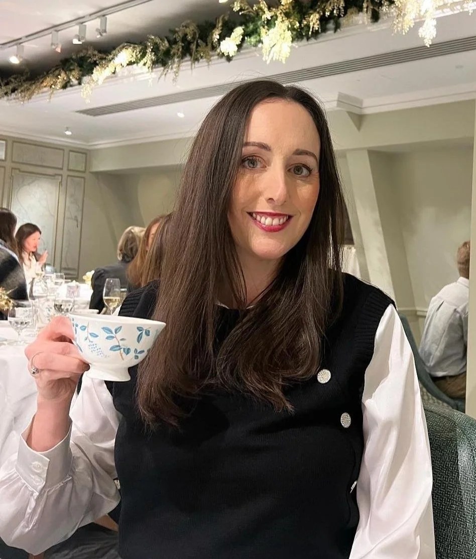 Karen McGovern Smart Hazardous Waste Solutions Finance Director, in a black sleeveless top over a white shirt is holding a floral teacup in a restaurant with people dining in the background.
