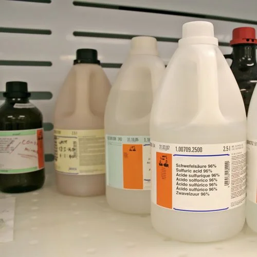 Plastic bottles containing sulfuric acid which is a hazardous waste, on a shelf in a laboratory.