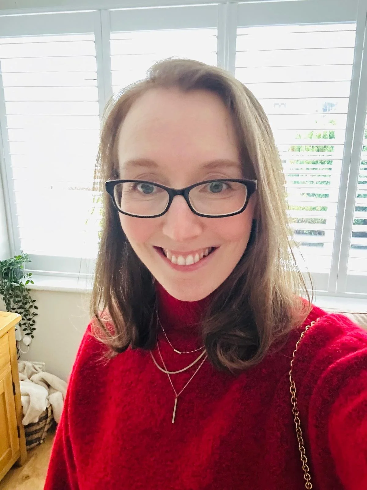 Person wearing glasses and a red sweater, smiling indoors with white shutters in the background.