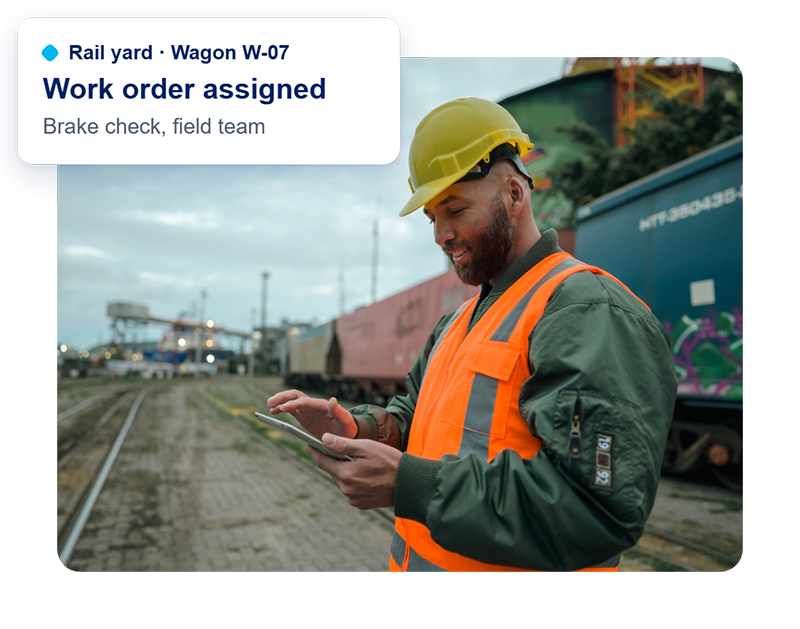 Rail yard worker in a hard hat and safety vest uses a tablet beside freight wagons. A card shows a work order assigned for Wagon W-07.