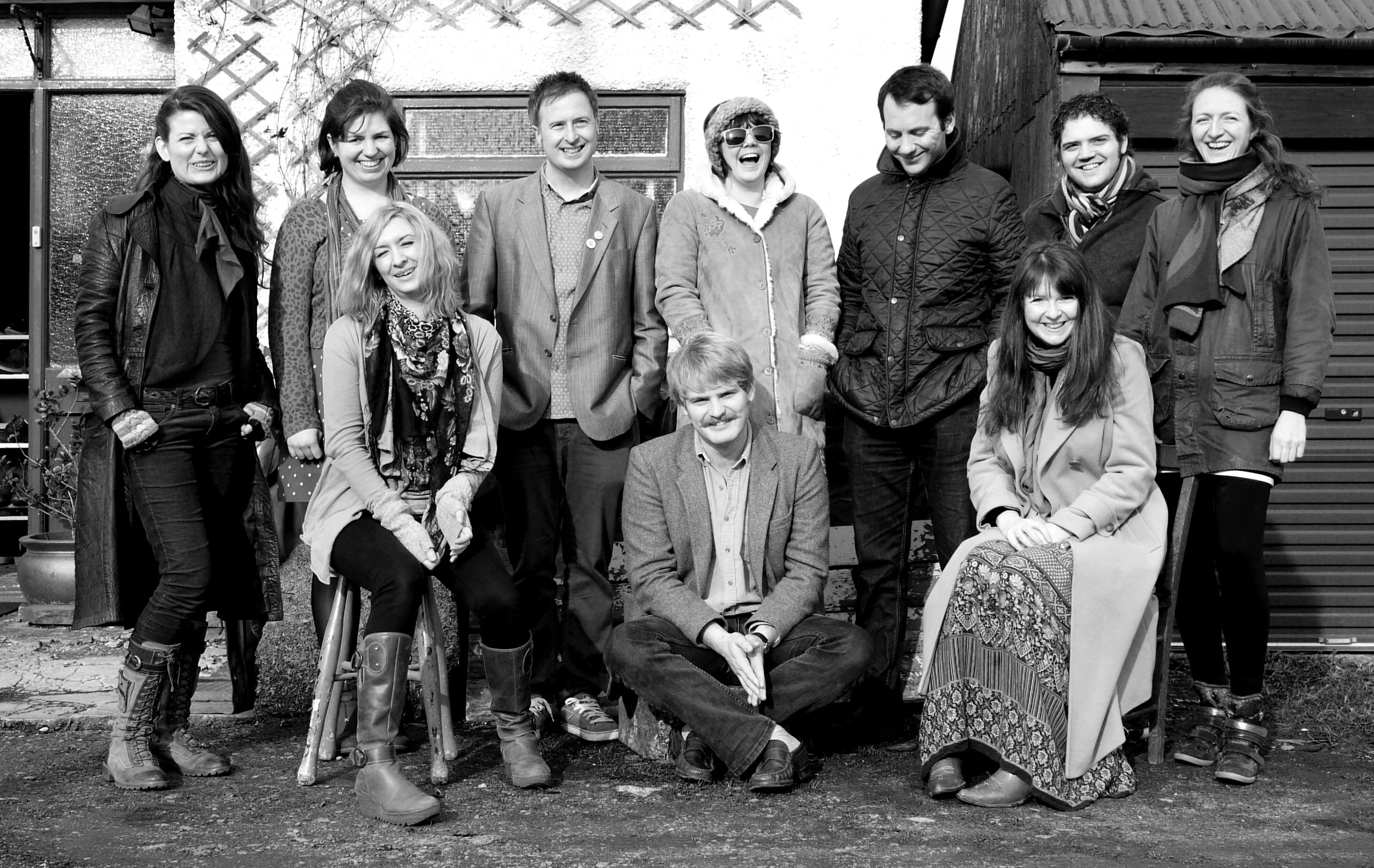 Group of ten musicians from 10 Mewn Bws, posing outside with varying clothing styles, including coats and scarves, in front of a rustic building.