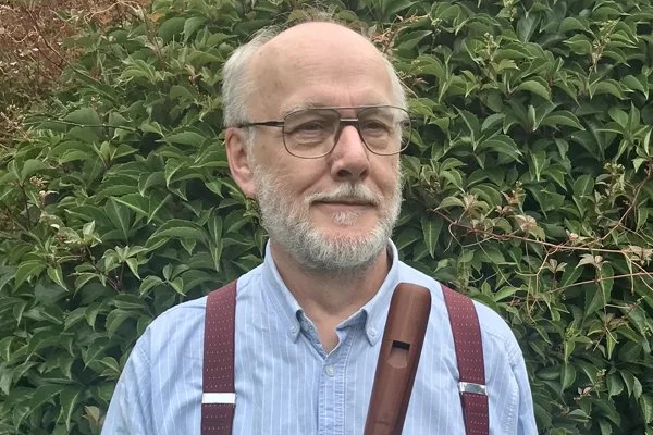 Dr Hugh Jones, holding a recorder and standing in front of green foliage.