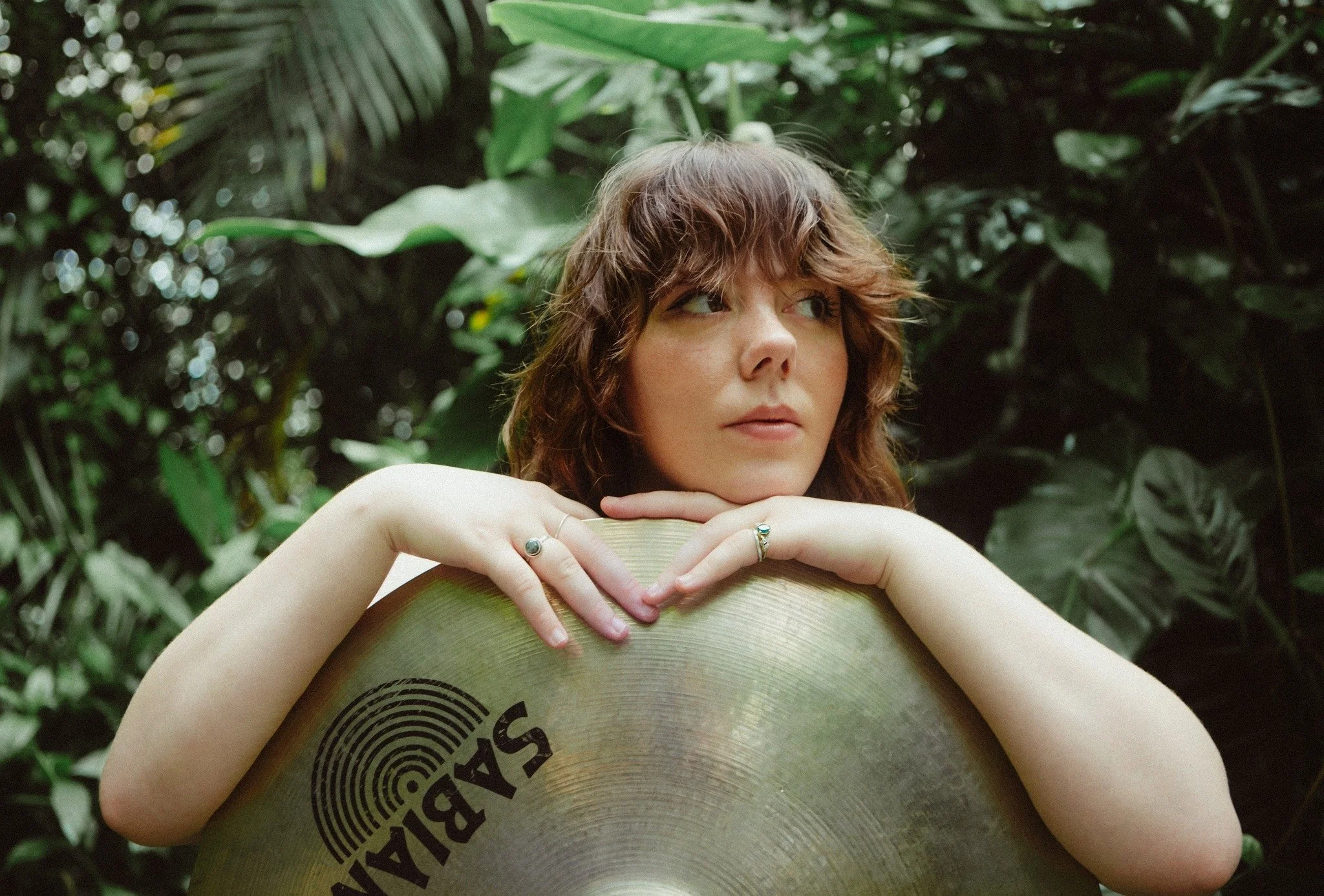 Meg Cox, posing with a cymbal in a lush, green forest setting.