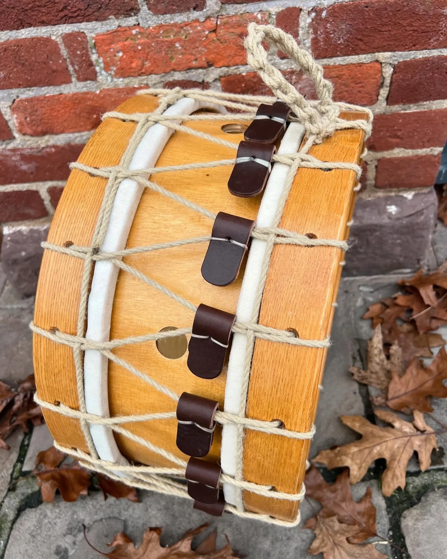 Wooden drum with rope tension, brown leather tabs, and autumn leaves on the ground, placed against a brick wall.