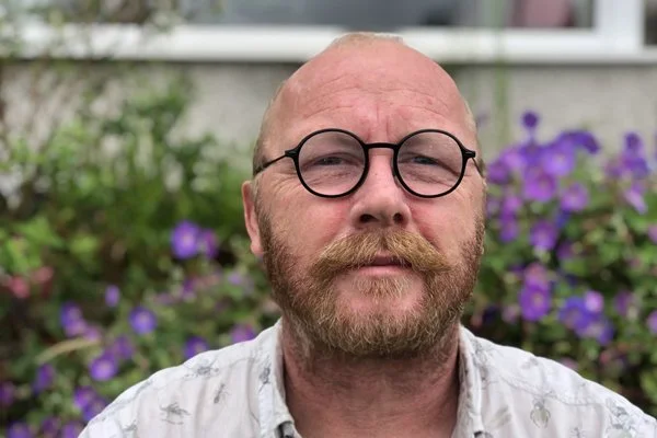 Danny KilBride, glasses and mustache, in a garden with purple flowers.