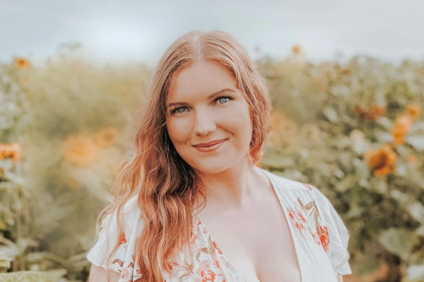 Gwenno Roberts, with long hair in a floral dress standing in a sunflower field.