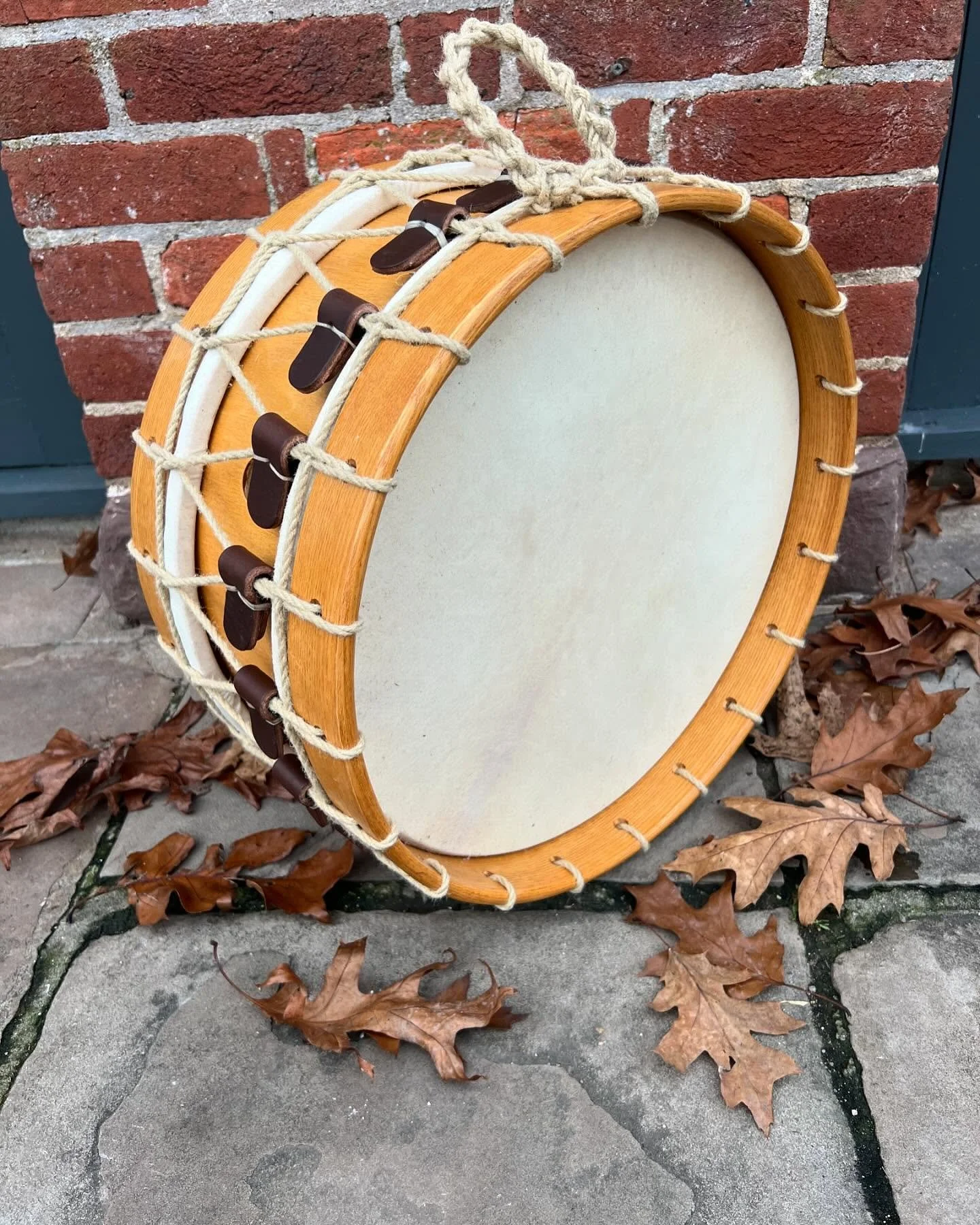Traditional drum with rope tension and wooden frame, resting against a brick wall with fallen leaves on the ground.