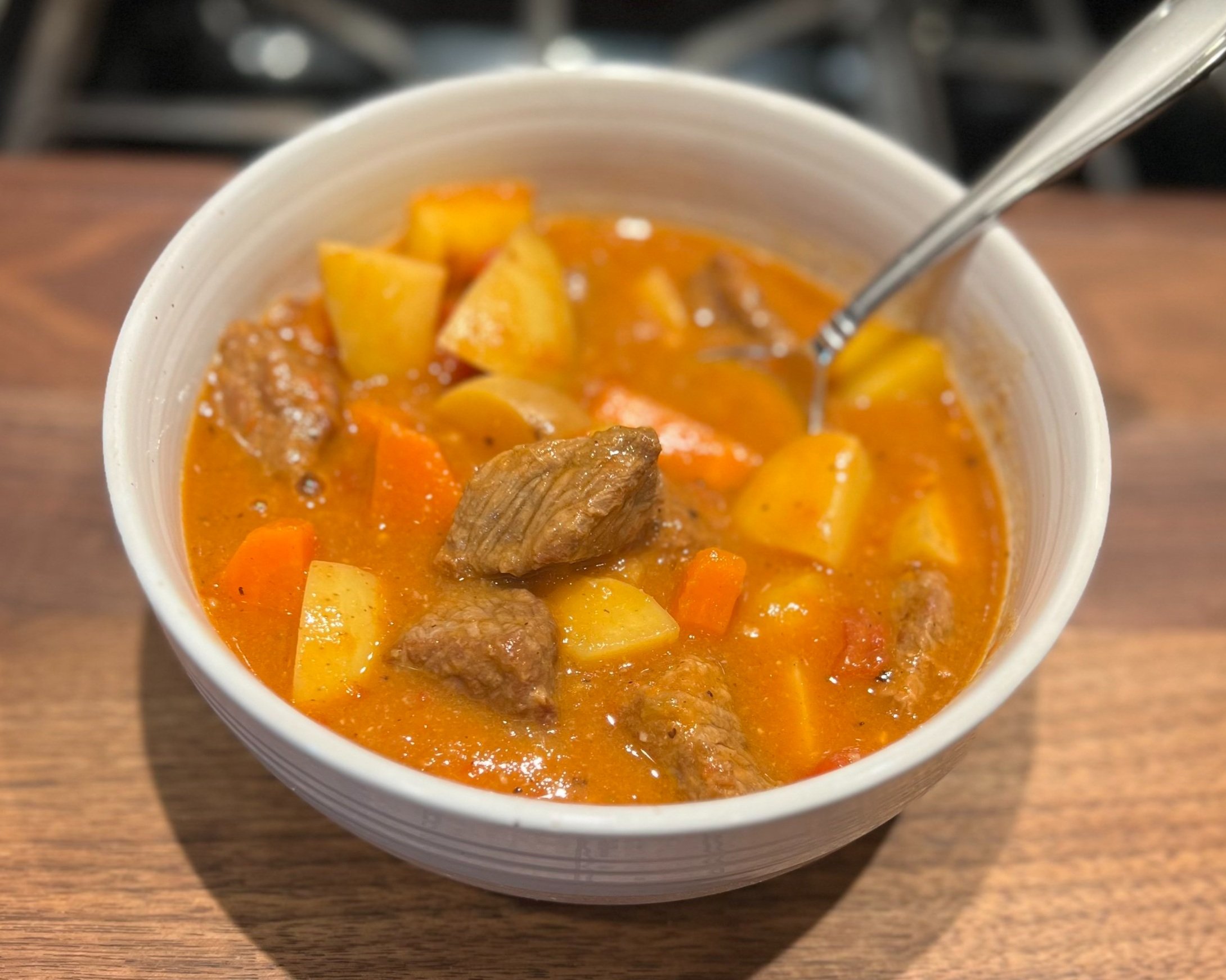 A bowl of beef stew with chunks of potatoes, carrots, and beef in a thick, orange-brown broth, with a spoon in the bowl.