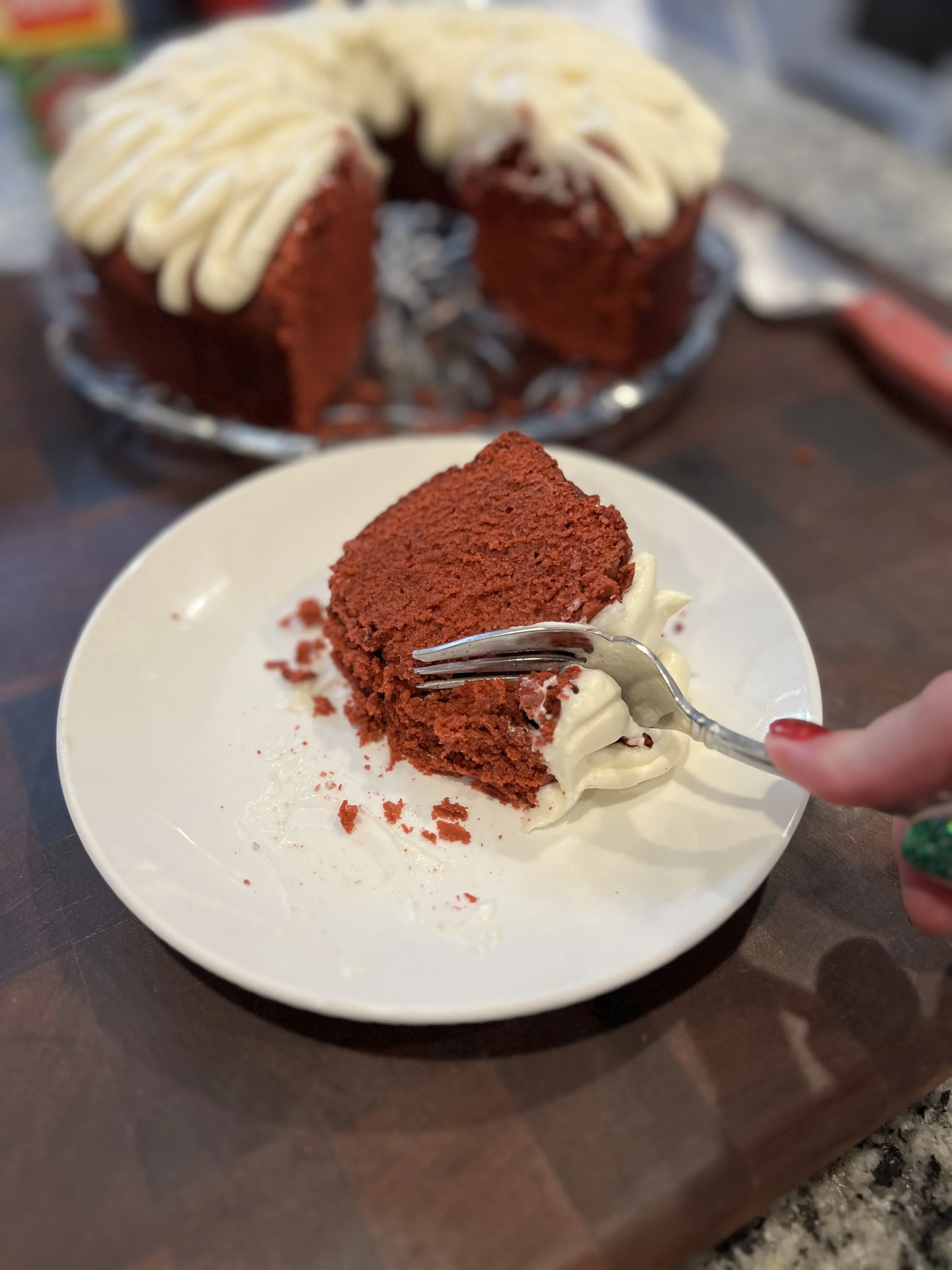 Slice of red velvet cake with cream cheese frosting on a white plate. A hand with a fork is ready to take a bite.