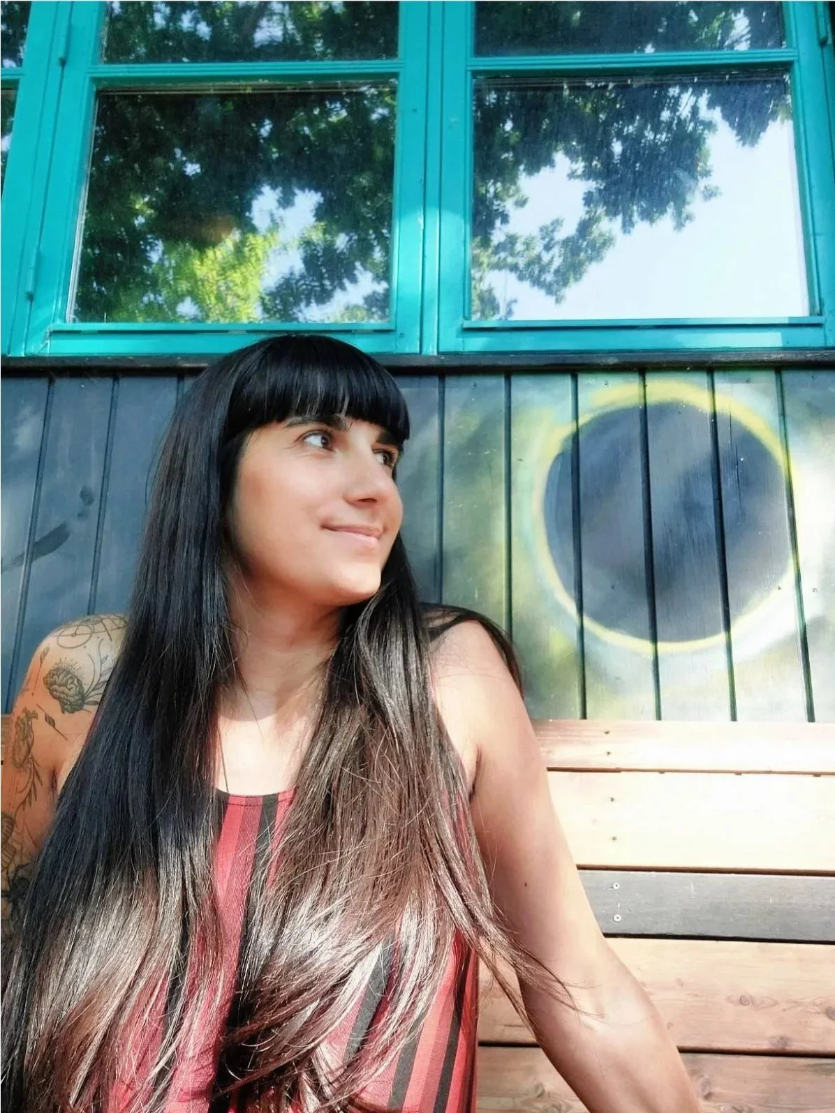 A woman with long dark hair and tattoos on her arm is sitting outside, looking to her right with a slight smile. Behind her is a painted wall featuring a crescent moon and circle, with large window panes above showing reflections of trees.