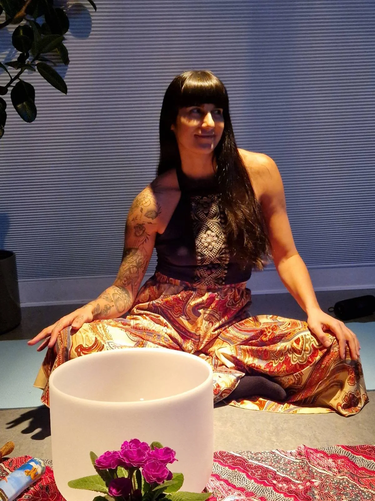 A woman with long black hair and tattoos on her left arm sitting cross-legged on a mat, next to a white singing bowl with a pink flower, in a room with a textured gray wall.