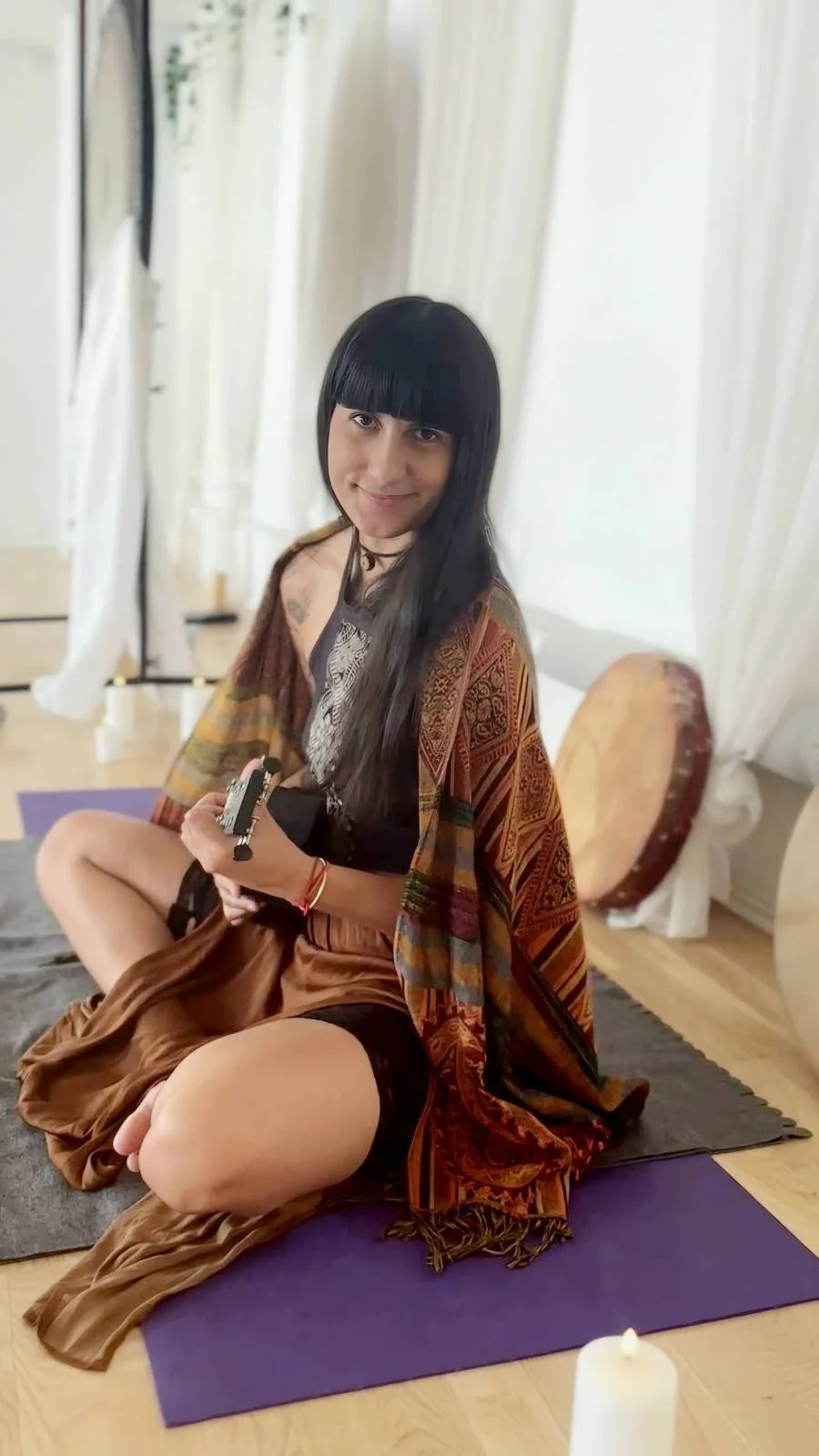 A woman with long black hair and bangs sitting cross-legged on a purple yoga mat, holding a set of keys, in a room with white curtains and a large drum in the background.