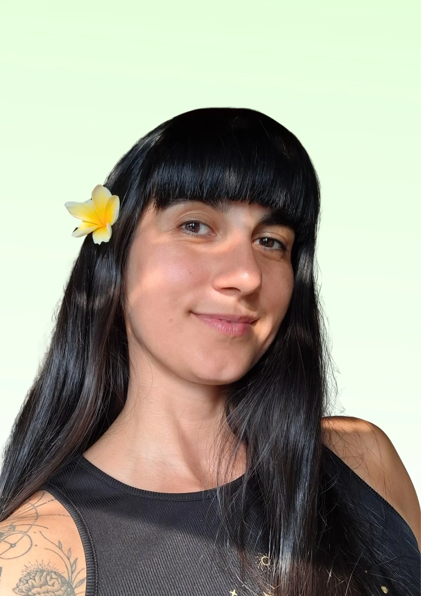 A woman with long black hair and bangs is smiling, wearing a black tank top, with a yellow flower tucked behind her ear, and showing a tattoo on her shoulder. The background is a plain light green color.